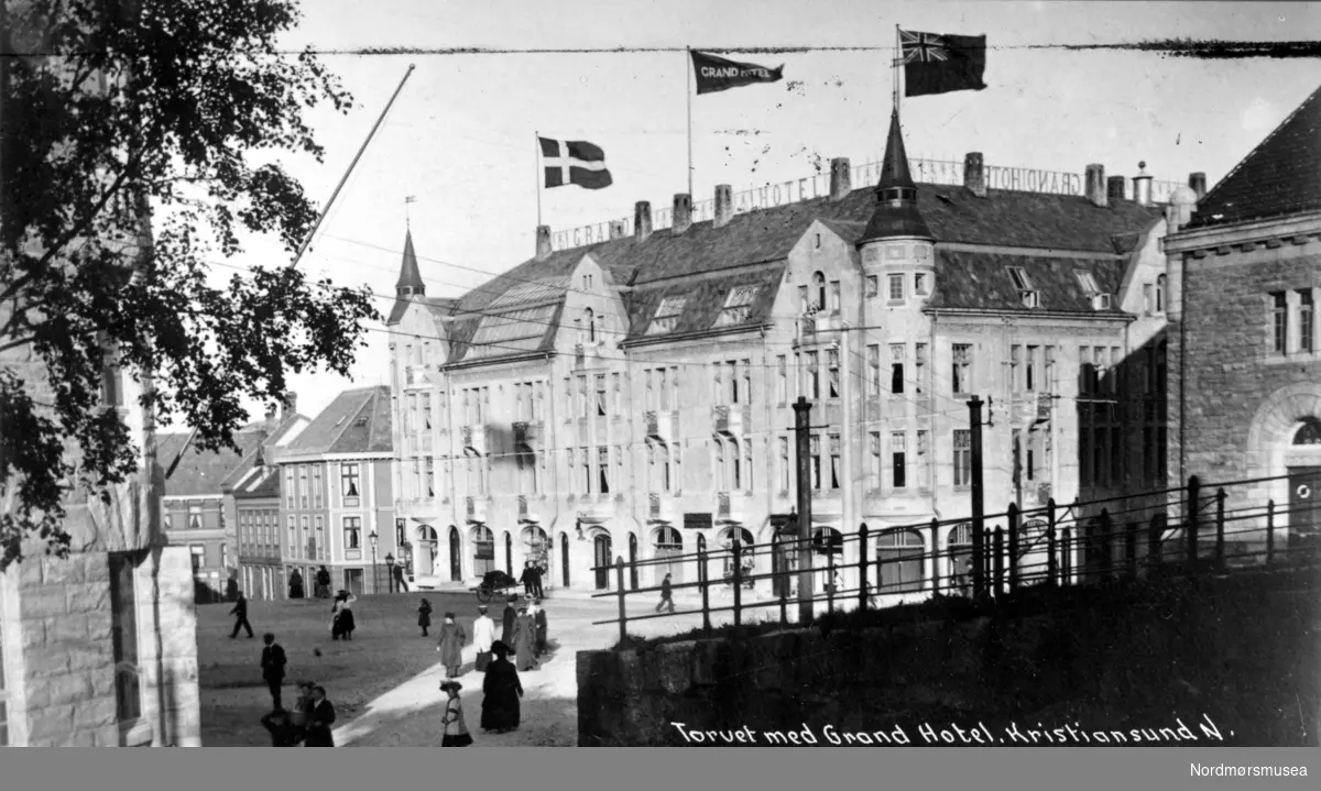 Foto fra begynnelsen av Parkveien som startet her fra Torvet på Kirkelandet i det gamle Kristiansund. Her ser vi i bakgrunnen fra venstre Grefsnes glass, det nylige oppførte Grand Hotell (nr. 2 i rekken) og Norges Bank. Byens første Grand Hotell - et flott sveitserbygg i tre - brant ned til grunnen, 7.nov. 1907 etter en brann i vedskjulet. Det ble besluttet at hotellet skulle bygges opp igjen, denne gang i Jugendstil, og arkitekten ble Kristen Tobias Rivertz. Hotellet stod ferdig januar 1909. Ellers så er gaten mellom Grand og Norges bank er Bernstorffstredet. Ellers så er det store murbygget vi ser litt av i front til venstre kommunehuset. Fra Nordmøre Museums fotosamlinger.