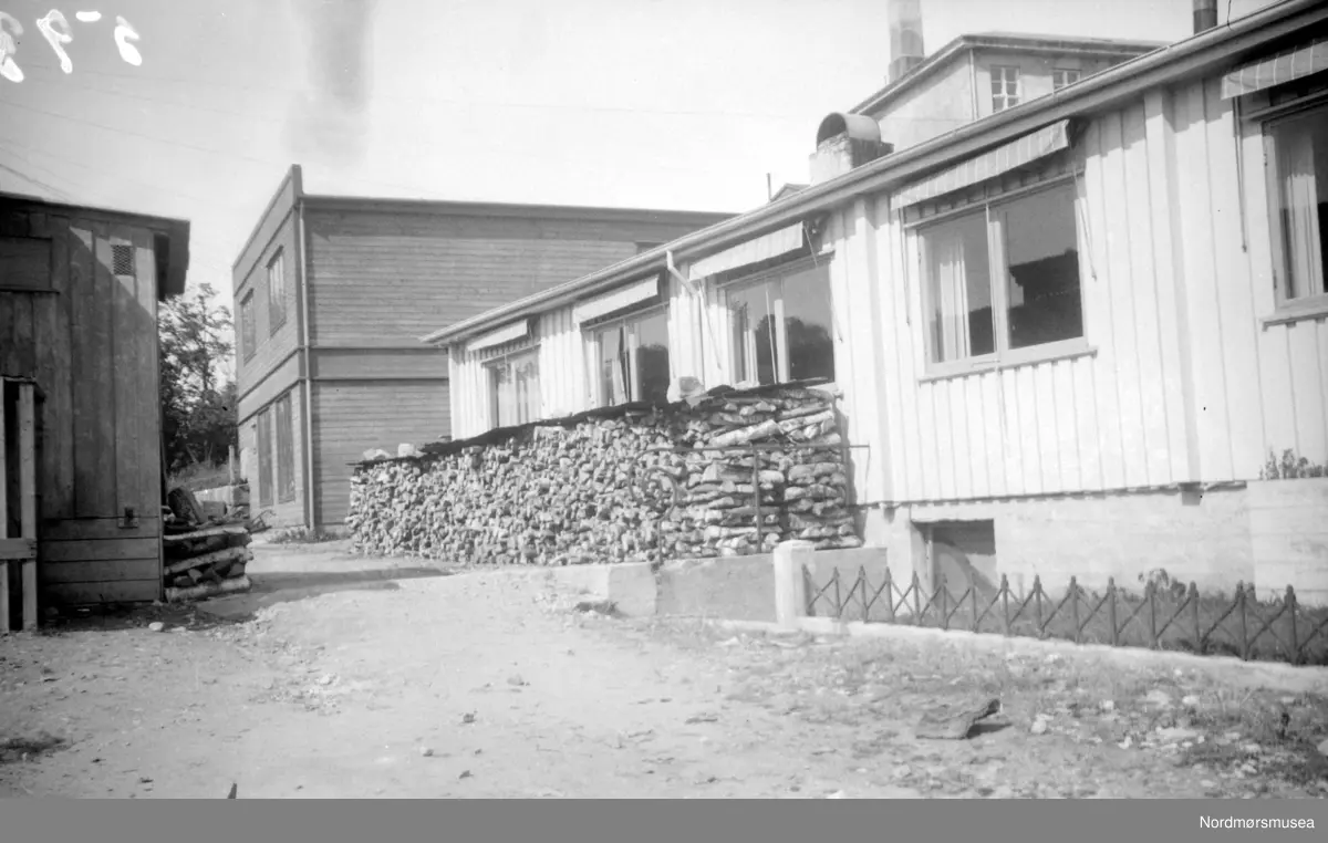 Utenfor Kristiansund og Nordmøres Forretningbanks brakke, juni 1943. Forsyning av vinterved stablet oppetter veggen. Brakka ligger her på Enggatens skole forhenværende tomt. I bakgrunnen til høyre ser vi taket til Kristiansund Offentlige Høgre Allmenskole (KOHAS), senere Langveien Ungdomsskole. Bildet er datert juni 1943. Nordmøre Museum.