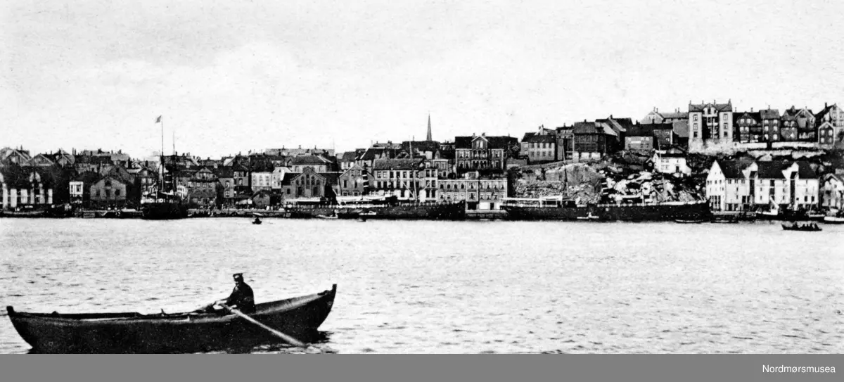 Foto fra havnebassenget i Kristiansund, med Kirkelandet i bakgrunnen. Vi ser også en robåt i forgrunnen. Postkortet er stemplet Kristian Kvande, Kristiansund N. og har innskripsjonen "Parti af Kristiansund N." Bildet er trolig fra begynnelsen av 1900 tallet. Fra Nordmøre Museums fotosamlinger.