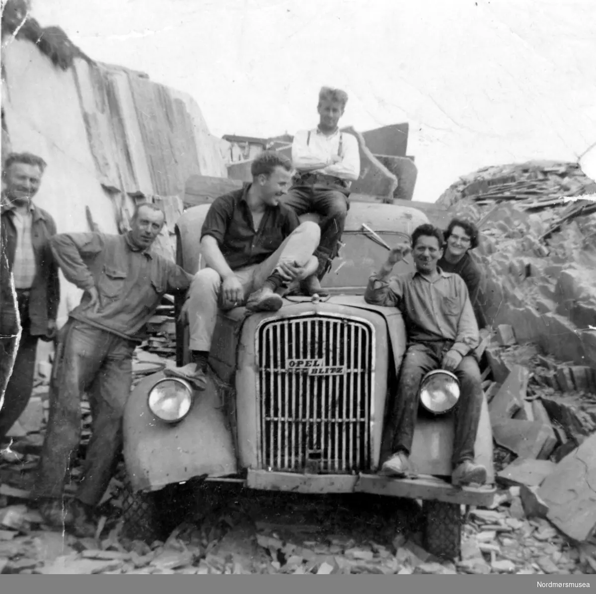 Foto av fem menn og en kvinne sittende på et gammelt vrak av en Opel Blitz lastebil, parkert i noe som minner om et steinbrudd. Det er ukjent hvem personene på lastebilen er og hvor og når bildet er tatt. Fra Nordmøre museums fotosamlinger.