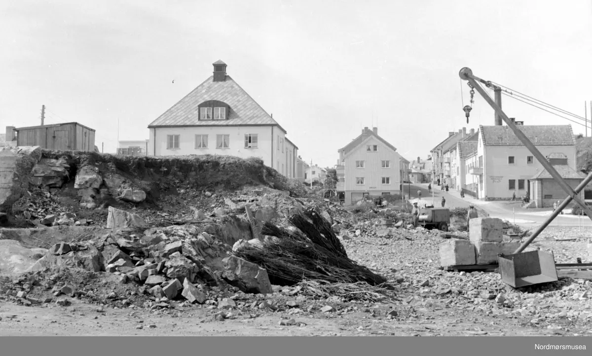 Siste rest av fjellhaugen der Arbeiderforeningen og Rådstua stod. I bakgrunnen ser vi rester av en tysk bunker som ble bygget under krigen 1940-45, og over denne bunkeren hadde da Arbeiderforeningen stått før bybrannen i de sene aprildagene 1940. Bak denne igjen ser vi det nye bygg for folkebiblioteket. I bakgrunnen ser vi Indremisjonsbygget og oppover Marstrands gate. Badet (seinere Nor-Pro) til høyre, revet (først parkering for ny-banken, pr 2007 boligblokk). Her holdt lastebilsentralen til i 1960-åra, pr 2007 Hydro bensinstasjon, og drosjene er fortsatt pr 2007 foran Misjonshuset. 
Inngangen støpt igjen i januar 2017 i forb. med butikk i Tinghuset. ( Regelbau 633 for M19 bombekaster) (info fra Bjørn Ove Angvik - 30.01.2017)
 

Nordmøre Museums fotosamling