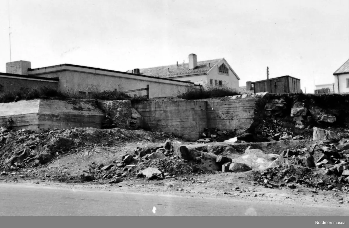 Siste rest av fjellhaugen der Arbeiderforeningen og Rådstua stod. I bakgrunnen ser vi rester av en tysk bunker som ble bygget under krigen 1940-45, og over denne bunkeren hadde da Arbeiderforeningen stått før bybrannen i de sene aprildagene 1940. Bak denne igjen ser vi det nye bygg for folkebiblioteket. I bakgrunnen ser vi Indremisjonsbygget og oppover Marstrands gate. Badet (seinere Nor-Pro) til høyre, revet (først parkering for ny-banken, pr 2007 boligblokk). Her holdt lastebilsentralen til i 1960-åra, pr 2007 Hydro bensinstasjon, og drosjene er fortsatt pr 2007 foran Misjonshuset. 
Inngangen støpt igjen i januar 2017 i forb. med butikk i Tinghuset. ( Regelbau 633 for M19 bombekaster) (info fra Bjørn Ove Angvik - 30.01.2017)
 

Nordmøre Museums fotosamling