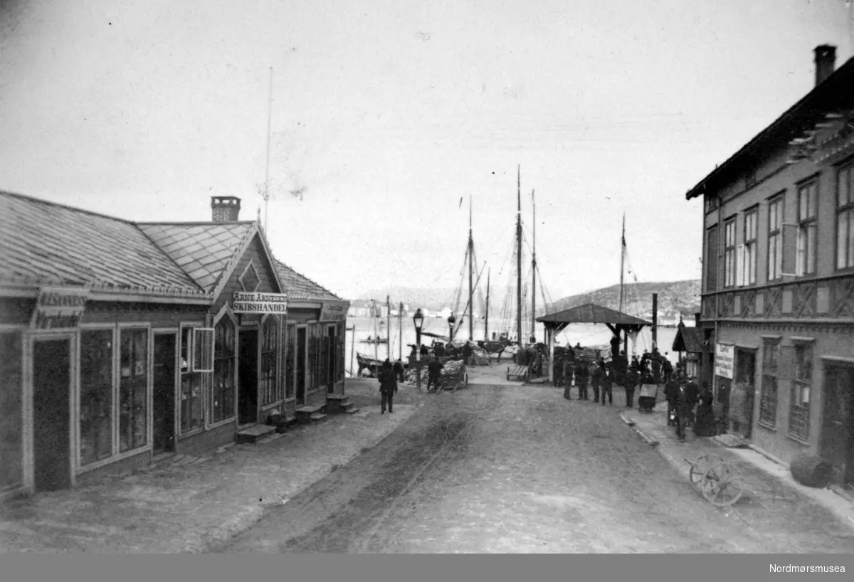 "Kirkealmenningen". Foto fra deler av bebyggelsen i Kirkeallmenningen på Kirkelandet i Kristiansund. Her ser vi blant annet Arne Arnesens Skipshandel til venstre på bildet, men Allmenningen i enden av gaten. Bildet er fra fotoalbumet "Christiansund i Fotografier". Bildet er trolig fra 1883, etter informasjon i Willhelm Lund-samlingen. Fra Nordmøre Museums fotosamlinger. 