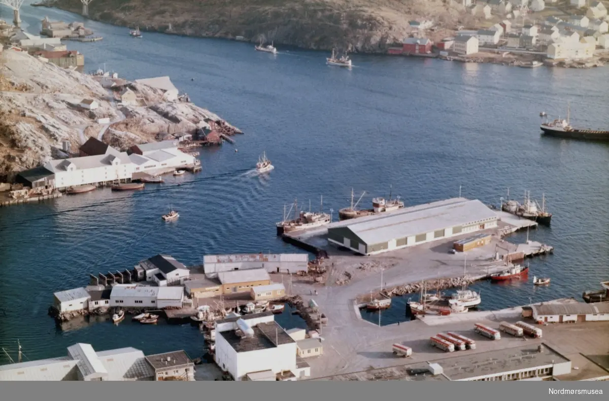 Flyfoto over havna i Kristiansund. Helt i forgrunnen kan vi se Rutebilstasjonen og Fryseriet på havna i Kristiansund. På Devoldholmen var det mange små virksomheter; sykkel/mopedverksted, fargehandel, verksteder. Tollbod. Over vågen kan vi se Milnbrygga med føringsbåter foran. datert 20 februar 1965. (Fra Nordmøre Museums fotosamlinger)