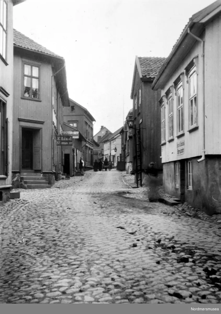 Gateparti fra Øvre Langgate i Tønsberg, hvor vi ser butikker langs en brosteinsgate. En av butikkene til venstre er ";E. Lind - Skomaker";. Endre Lind drev stort skomakerverksted fra 1875 på hjørnet av Øvre Langgate og Prestegaten (Tønsberg Historie(?) s. 178). I bakgrunnen ser vi personer med hest og kjerre. Fra Nordmøre Museums fotosamlinger. /Reg:EFR2013/



