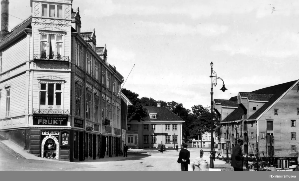 Johnsenhuken, med Ottesengården til venstre og med Johnsengården midt på bildet i enden av gaten. Johnsengården ble også brukt til Børslokale. Til høyre på bildet finner vi Johnsenbryggen. Fruktbutikken på hjørnet av Ottesengården tilhører M. Endresen. Børs, Børslokale, tannlege, bygård, kjøpmannsgård, mennesker, menn, bygninger, hus, brygge, kai,
 Fra Nordmøre Museums fotosamlinger.
