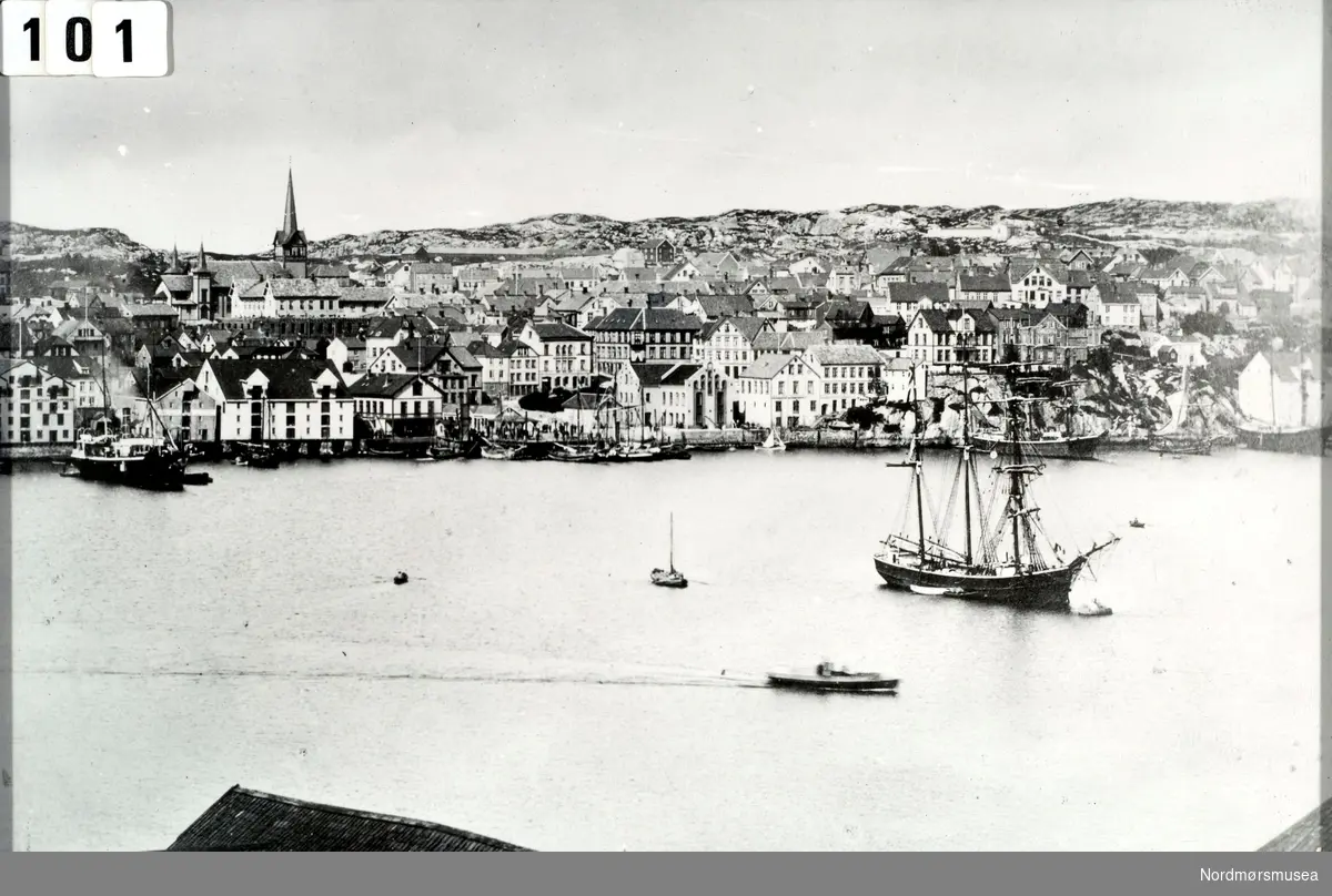 Foto over havnebassenget i Kristiansund, med et oppankret seilskip ute i bassenget. I bakgrunnen ser vi Kirkelandet, med Kirkelandet kirke til venstre. Bildet er sett fra Nordlandet, og er datert til 1885. Fra Nordmøre Museums fotosamlinger.
