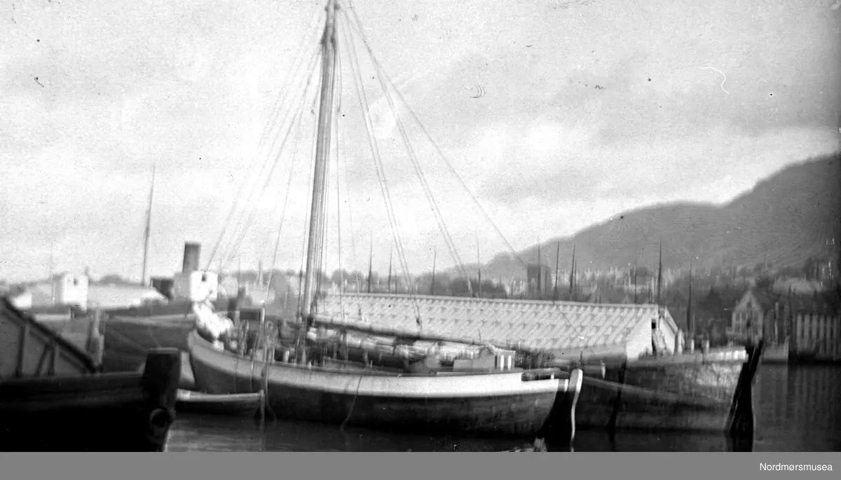 Båten i forgrunnen er en seilskøyte. Den som ligger bak kalles i Kristiansund en lekter (for klippfisk); dvs en føringsbåt. "Teltet" er et overbygg på lekteren, rigget slik ble de vanligvis kalt lossementsbåt  og ble brukt som flytende bolig for fiskere eller lager. Fjellene i bakgrunnen gjør det usikkert om det kan være i Kristiansund. Det gjør at det blir vanskelig å si eksakt hva fartøyet burde kalles og hva det har vært brukt til. ca 1900. Fra Nordmøre Museum sin fotosamling.