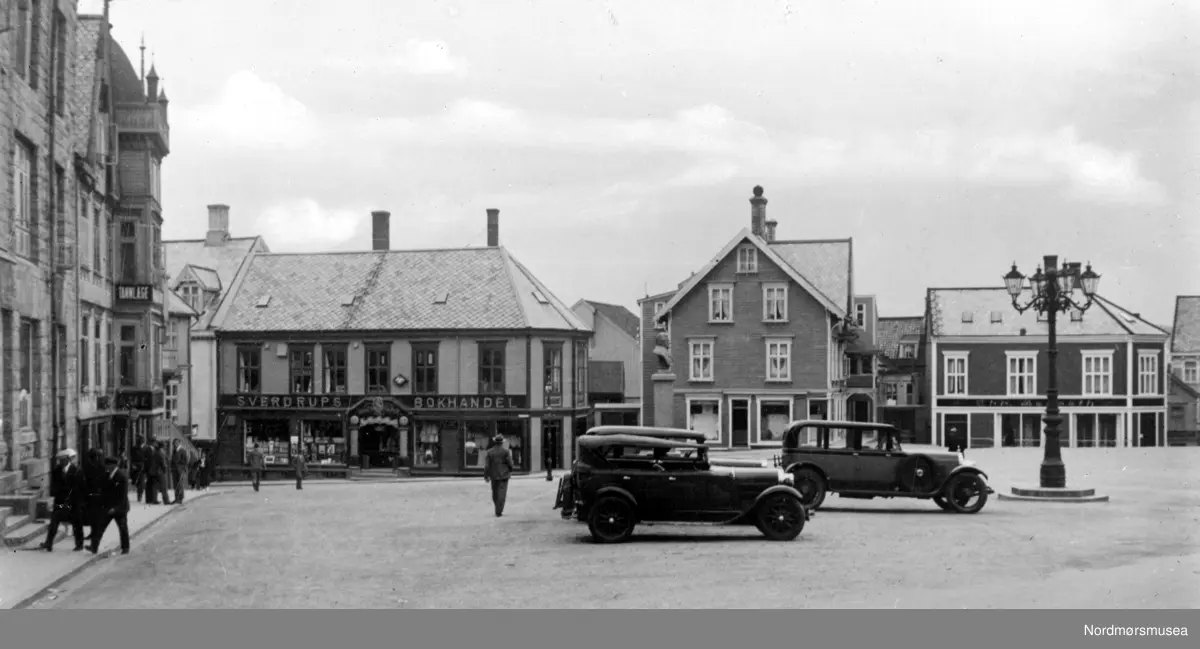 På bildet ser vi fra det gamle Torget på Kirkelandet i Kristiansund. De fleste av disse bygningene brant ned under bybrannen 28. april 1940, da tyskerne bombet byen. I bygningen helt ytterst til venstre ser vi litt av Kommunebygningen. Deretter kommer en bygning som huset blant annet Tannlege Helsing og Kafe Kristiansund. Deretter kommer Sverdrups Bokhandel som lå i Torvgaten 9. Georg Sverdrup hadde kjøpt lokalene 1. oktober 1930, fra Fosna Mållag, som reiste bygget i 1910. Bedriften var da eid av G. Sverdrup. 28. april 1940 brant butikken ned til grunnen og den 24. september 1940 ble branntomten overført fra G. Sverdrup til Sverdrups Bokhandel A/S. Bygget i midten er Dahls bygård og inneholdte blant annet en konfeksjonsforretning, mens huset ytterst til høyre er Berseth, som inneholder blandt annet manufaktur og teppeforetning. Grefsnes Glass kommer igjen til høyre for Berseth, men den forretningen er ikke med på dette bildet. På torgplassen foran kommunehuset finner vi den svært så kjente ";Lysekrona";, en smijern- gatelykt som er smidd ved en av byens skipsverft. Denne står foran Festiviteten per 2007. Nordmøre Museums bildesamling
