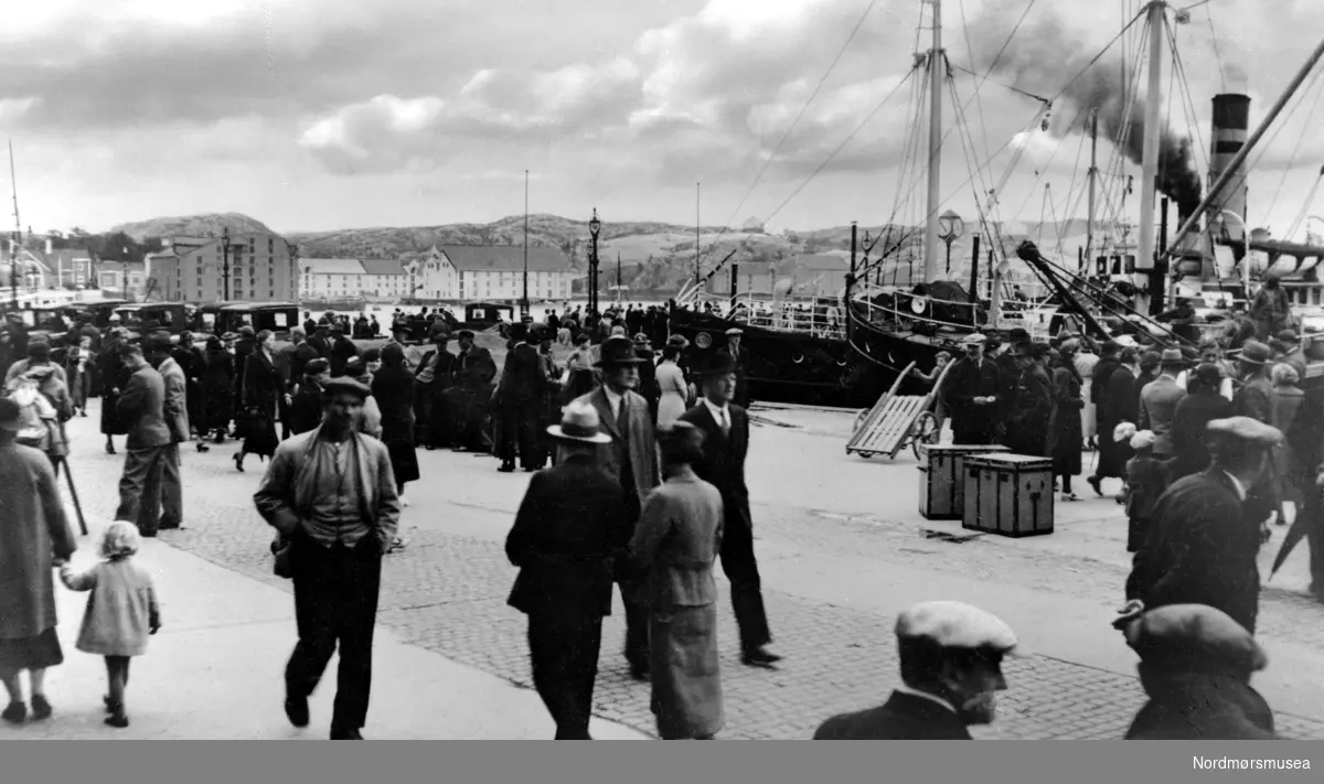 Foto fra Kirkelandet allmenning/Piren og Dampskipkaia, på Kirkelandet i Kristiansund kommune, hvor vi ser fullt av folk i gatene, med biler i bakgrunnen og dampskip til kai. Fotograf er trolig Georg Sverdrup, og datering er sannsynligvis fra perioden 1930 til 1939. Fra Nordmøre Museums fotosamlinger.