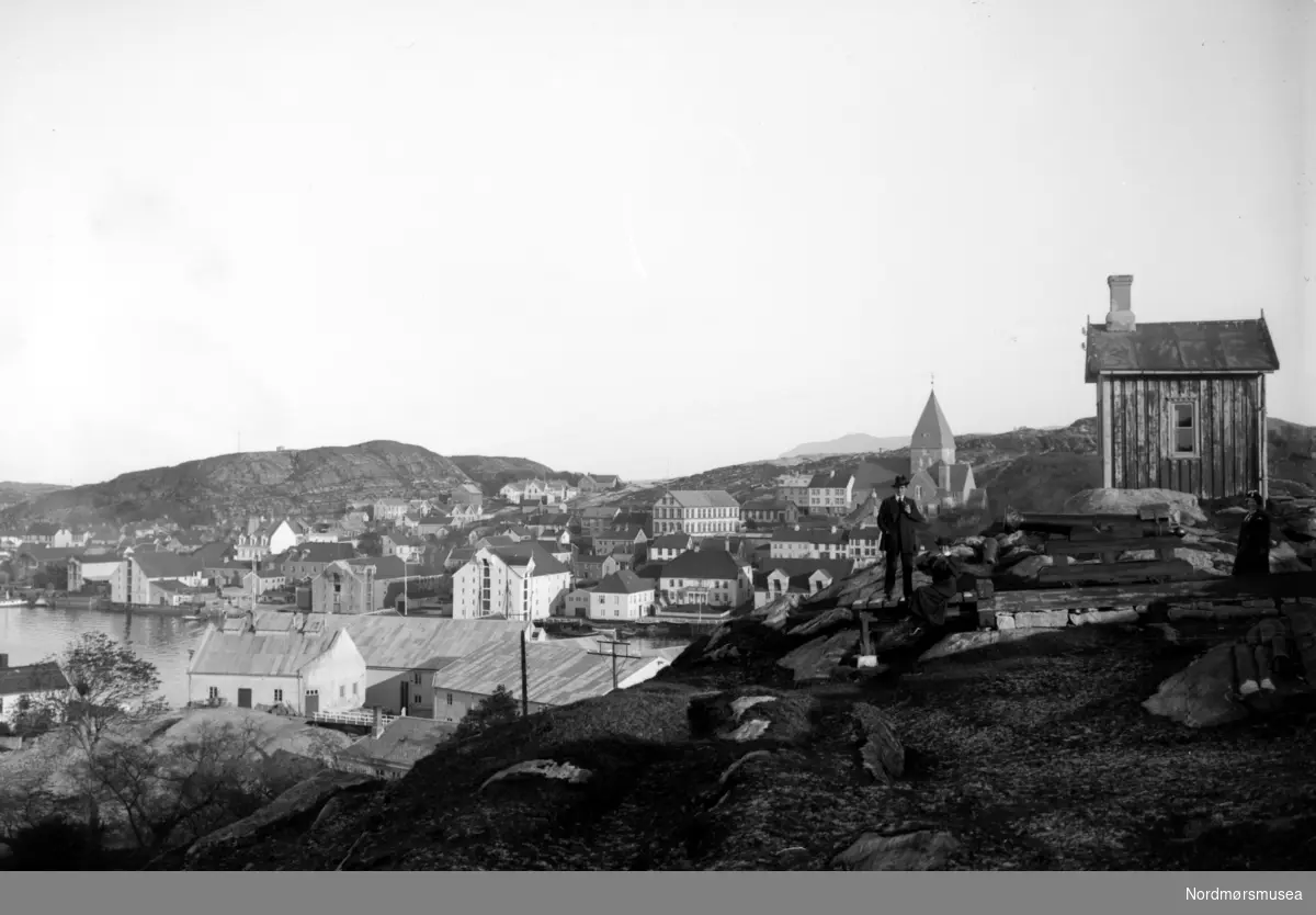 På bildet ser vi en mann oppstillt foran en enkel trehytte. I bakgrunnen ser vi over til Nordlandet, hvor Nordlandet kirke, med Nordlandet skole til venstre for kirken koomer klart frem i bildet. Fra Nordmøre Museums
fotosamlinger.
