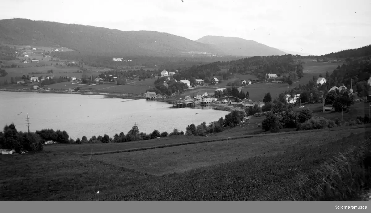 Foto muligens fra Tingvollfjorden i Tingvoll kommune, hvor vi ser bygda omkranse sjølinjen og strekke innover mot åsene der. Fra Nordmøre museums fotosamlinger. 