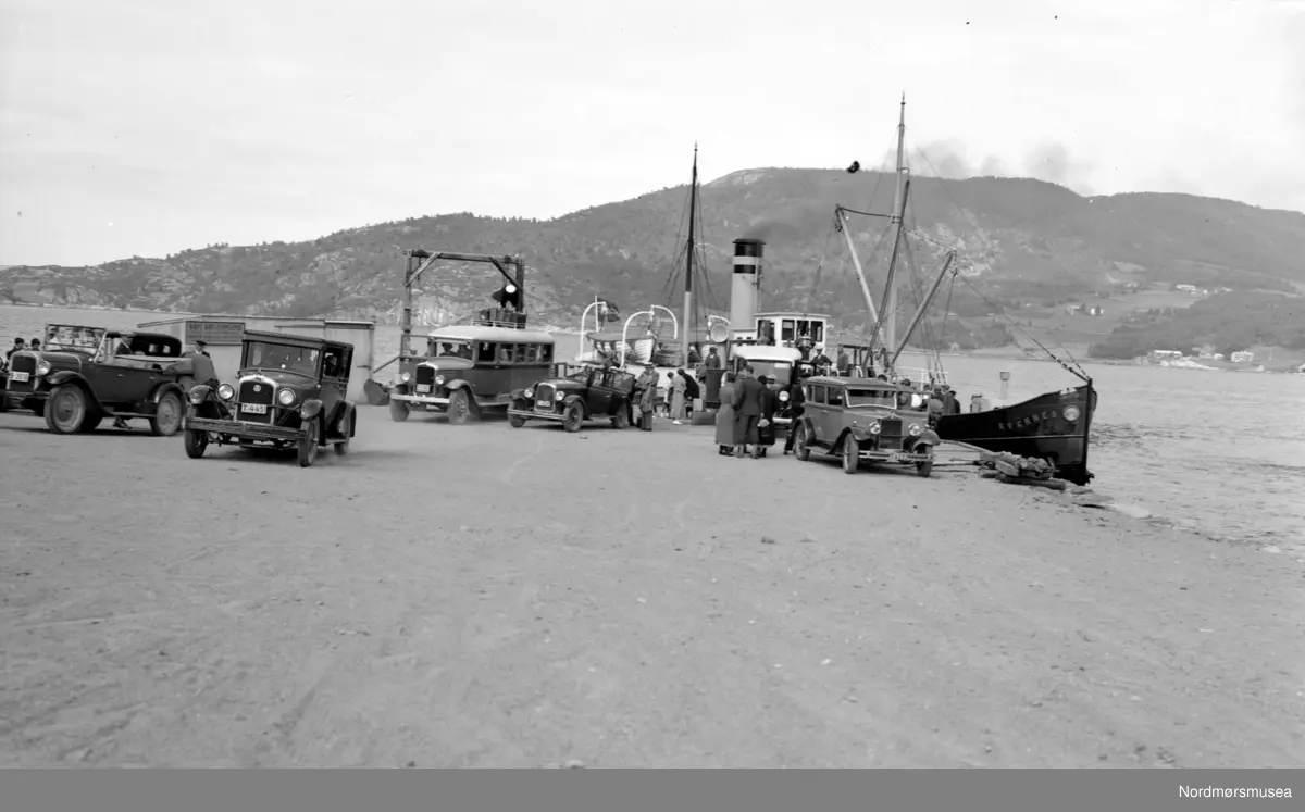 Rutebåt DS Kvernes ved Gjemnes fergekai. Flere biler og en buss står parkert foran. Bilen til venstre er en Chevrolet 1927, bussen muligens en GMC. (Info: Ivar E. Stav 2016). Fotograf er G. Sverdrup.  Dette bildet kan tidfestes nærmere, til 1932-1938. I 1932 ble veien Batnfjordsøra-Gjemnes åpnet. Gjemnes fikk båtanløp fra Kristiansund, og trafikken til Batnfjordsøra kunne gå med bil eller buss. Noen rutebåter, som D/S Kvernes på bildet, kunne ta med noen få biler. Først i 1938 fikk Gjemnes fast ferjesamband med B/F Trygge som ble satt i rute Kristiansund-Gjemnes-Torvikbukt. Dette bildet er ellers gjengitt og omtalt på side 294 i Ulstein, R. (1995). "Som skyttel i vev" (bind 1). Molde: Møre og Romsdal fylkesbåtar. (Info: Sveinung Berild - 09.11.2016). Fra Nordmøre museums fotosamlinger. Samme bilde som Kmb-1987-005.3953
