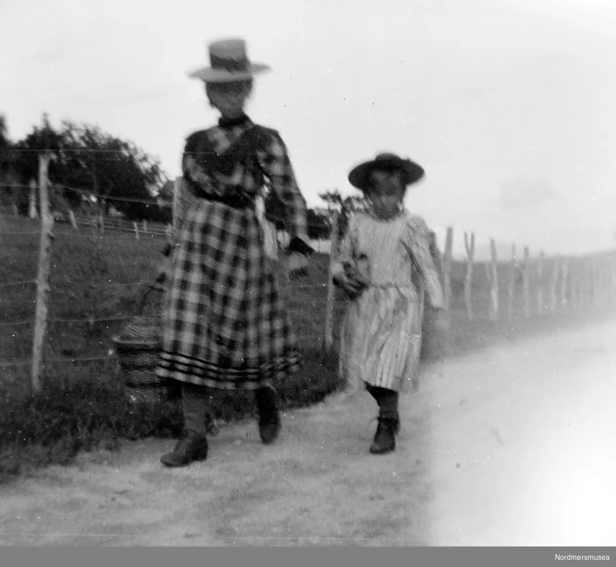 Uklart foto av Margrethe Barman Sverdrup og en liten pike langs en landevei/grusvei, med en piggtrådgjerde og beitemark like ved. Fra Nordmøre Museums fotosamlinger.
