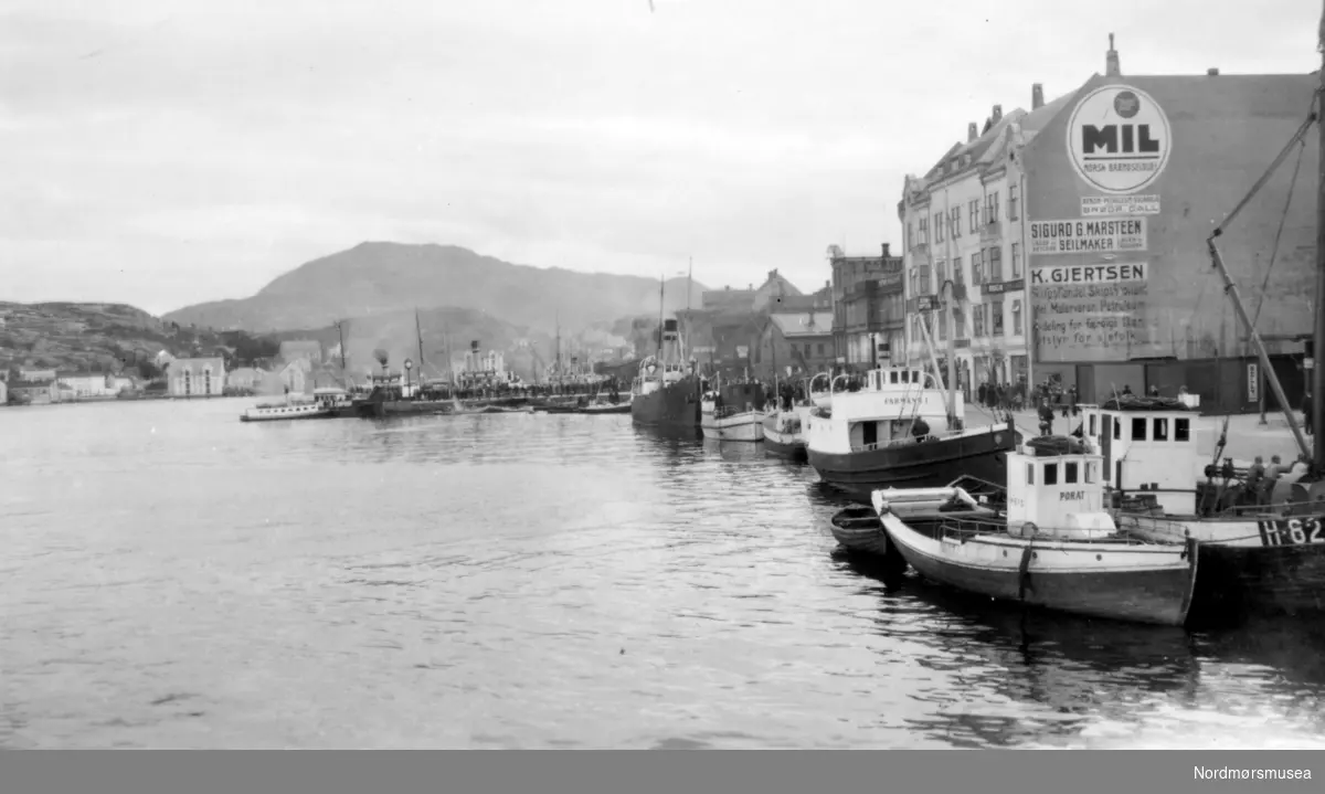 Et bilde sett fra havna, og langs Vågeveien med alle fiskebåtene, med Piren stikkende ut i front. Her ser vi sundbåten liggende ved kai. Den går i trafikk mellom øyene i Kristiansund. Serie, se samme motiv med hurtigruta på. Fra Nordmøre Museum sin fotosamling
