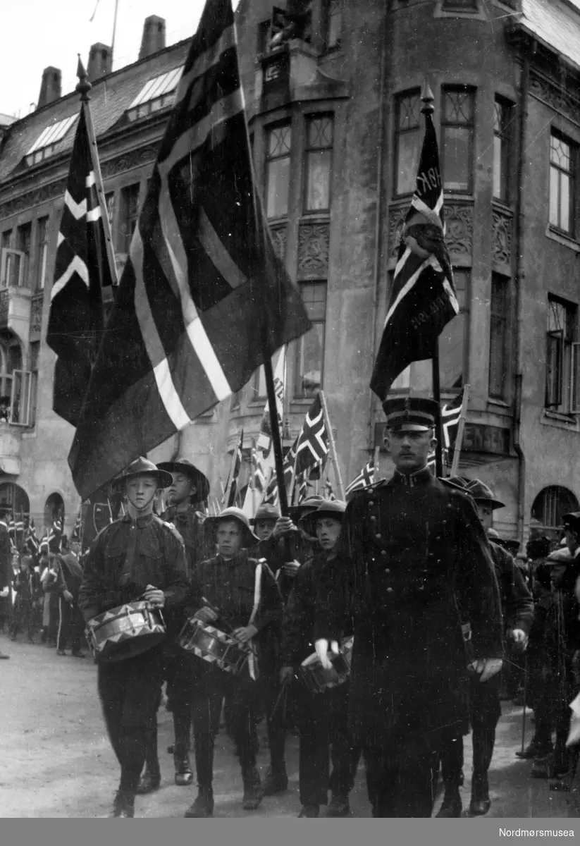 17. mai-togets flaggborg sammen med  speidernes trommeslagere fremst i toget utenfor Grand Hotel på Torvet, Kirkelandet, Kristiansund.  Politikonstabelen først i toget kan muligens være Bang eller Johan Eikrem.  Sistnevnte var den første leder for guttespeiderne da Norsk Speidergutt-Forbund ble stiftet i 1911, og det var mye takket være han at oppslutningen ble så god i Kristiansund.  Han ble senere leder for speiderne i hele fylket.   På denne tiden var det stor konkurranse guttekorpsene i mellom og de konkurerte med trommer og masjering så snart en anledning bød seg.  Her er det speiderne som er først i toget.
Pikespeiderne kom med i 1920-årene. 

(Fra Nordmøre Museums fotosamlinger)