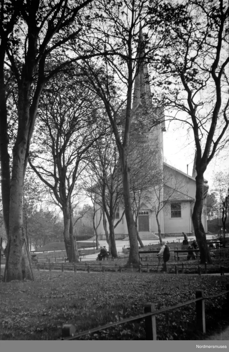 Kirkelandet kirke på Lyhshaugen, Kirkelandet i Kristiansund. Her med deler av parken i forgrunnen.  Etter forordning av statsmaktene 1858 skulle kommunene heretter ha ansvar for kirkebyggene, og kirkesaken ble et livlig diskusjonsemne i bystyre, presse og blant folk ellers gjennom flere år.  I 1875 vedtok bystyret å bygge ny kirke på Kirkelandet og flytte den gamle kirken over på Nordlandet, en bydel som i mange år hadde forlangt egen kirke. Tomten for den nye kirken ble Lyshaugen, oppkalt etter kjøpmann Lyhs, men et stort mindretall stemte for den gamle kirketomten på Torvet. Spørsmålet om kirkens plass hadde nemlig vært et nesten lidenskapelig diskusjonsemne i forveien, så vel offentlig som privat.  Bystyret vedtok at arkitekt Thrap-Meyers utkast skulle legges til grunn for nykirken. Et treskipet trekirke i Pseudo-gotisk stil og i motestilen de årene. Kostnaden var beregnet til 115.000 kroner, men da den var ferdig i 1878, var det gått med 160.000 kroner.  Kirken ble innvidd av biskop Grimelund, 18. september 1878. Den siste gudstjenesten i den gamle kirken ble holdt 15. september 1878 av res. Kap. Søholt. Ludvig Brinchmann var sokneprest og utnevnt året før.  På grunn av den store overskridelsen av byggesummen, og ellers de store utgiftene kommunen hadde i de årene med vannverket, skolebygg med mere, måtte vedtaket om flytting av den gamle kirken til Nordlandet annulleres. Den ble derfor solgt ved auksjon i begynnelsen av 1884 til byggmester Arnt Rønning og tre bønder på Kirkelandet, P. Glærum i Dalen, O. Flatvad i Brunsvika og Eide i Kaasbølmyra. De rev kirken og brukte materialene til nybygg i byen og til utbedring av uthus på gårdsbrukene.  Kallsboken for Kristiansund Prestegjeld forteller at den siste gudstjenesten ble holdt i Kirkelandet kirke søndag 21. april 1940. Det ble da holdt høymesse ved stiftskapellan J. S. Garpestad og aftensang ved tredjeprest H. G. Seim. Mandag 29. april gikk flyalarmen tidlig og tyske fly kom inn over byen. Ved 11:30 tiden ble det kastet sprengbomber og store mengder av brannbomber. Hele strøket oppover Haugan, Løkka og Vestre bydel ble da satt i brann. Kirkelandet kirke sto lenge urørt men om ettermiddagen ble også den antent og brant ned. Da kirken var truet av gnister allerede søndag kveld den 28. april ble det meste av altersølvet ført i sikkerhet: 90 særkalker, en vinkanne, en kalk, en disk og en brødeske. Videre ble dåpskanne og dåpsfat, to messehakler, den ene fra 1761 og den andre fra 1938 ført i sikkerhet. Resten brant opp dagen etter i brannen.   Etter krigen kom spørsmålet opp om gjenreisning av byens hovedkirke, og 24. mai 1964 kunne byen feire åpningen av nye Kirkelandet kirke. Denne ligger ved Roligheten.  Fra Nordmøre Museums fotosamlinger.  Kilde: Kirkelandets kirke. Festskrift ved kirkeinnvielsen 24. mai 1964. Reg: EFR

