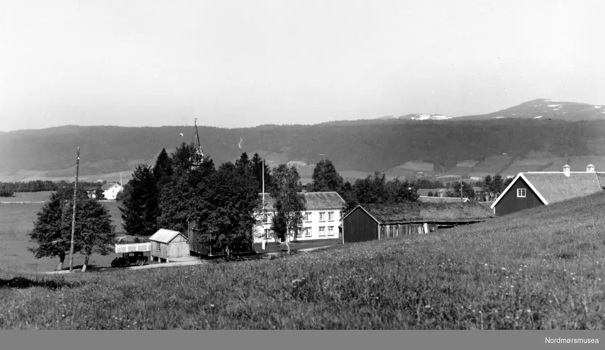 Foto fra Rindal prestegård i Rindal kommune. Her ser vi gården i front, mens lenger i bakgrunnen såvidt kan skimte kirketårnet bak trærne. Fotograf og datering er trolig Georg Sverdrup og datering er sannsynligvis fra perioden 1930 til 1939. Fra Nordmøre Museums fotosamlinger. Reg: EFR
