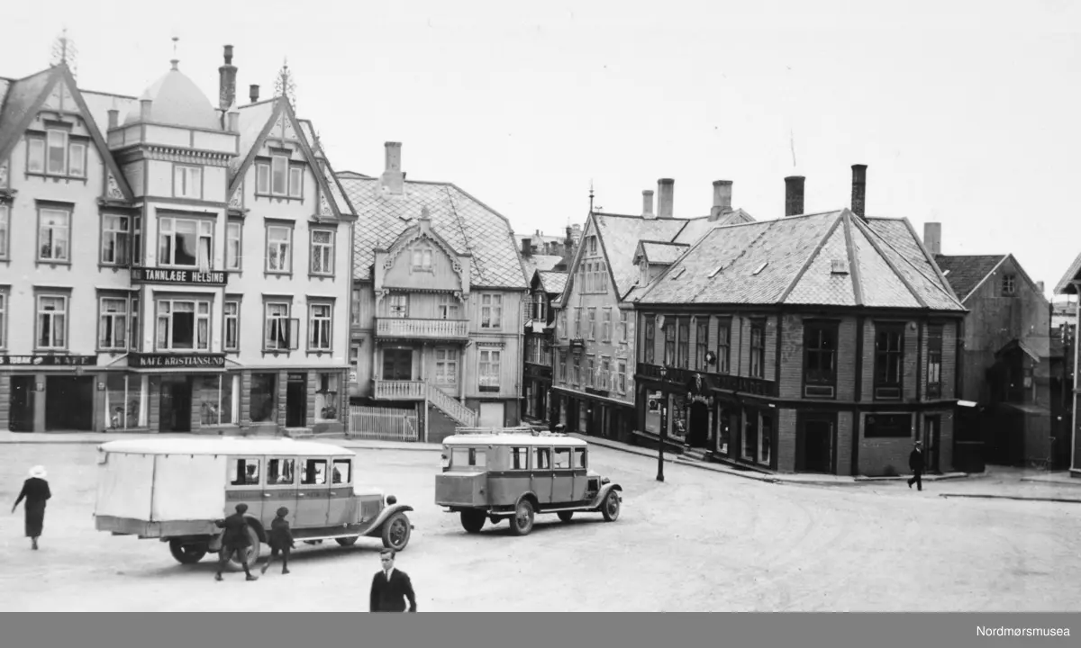 To busser står parkert på Torget på Kirkelandet i Kristiansund. Vi ser Sverdrups bokhandel til høyre i bildet. På bilen til venstre kan vi skimte Kristiansund-Oppdal Auto (KOA) på stripa under vinduene. Her er Oppdal stavet med P. Dette er nok av de tidlige rutebilene fra KOA. I 1932 åpnet ferjesambandet Kristiansund-Kvisvik. KOA og Surnadal Billag sine rutebiler kunne dermed følge ferja til Kristiansund sentrum. Det var først i 1938 at rutebilene fra Molde og Gjemnes kunne følge ferja til Kristiansund. Det var dette året ferjesambandet til Gjemnes og Torvikbukt åpnet. Bildet er derfor tidligst fra 1932. (info Sveinung Berild - 11.11.2016) Fra Nordmøre Museums fotosamling.