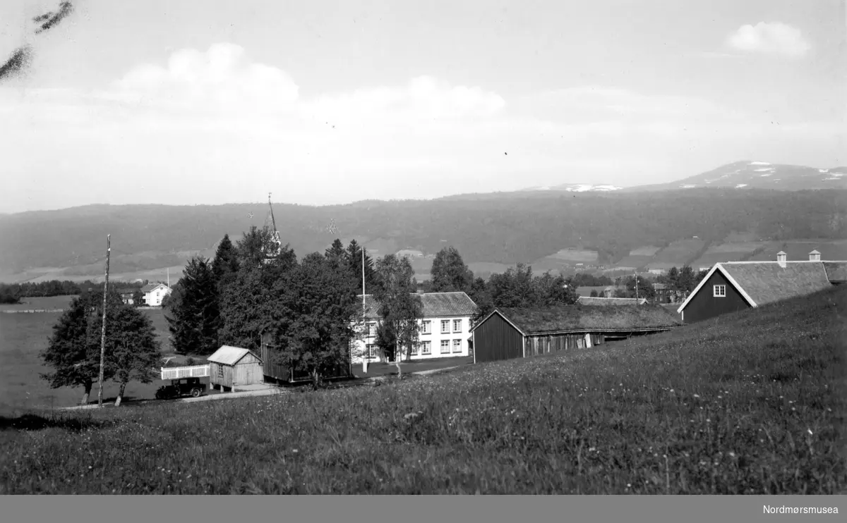 ";Rindal prestegård. Eneret G. Sverdrup 1935. 656."; Foto fra Rindal prestegård, med gårdshus i front og kirken like i bakgrunnen. Kirken er en korsformet kirke fra 1874. Fra Nordmøre museums fotosamlinger. Reg: EFR
