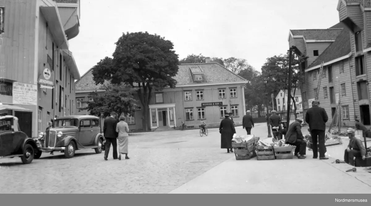 Foto av Johnsengården i Vågen på Kirkelandet i Kristiansund, her sentralt på bildet i bakgrunnen, med Johnsenbrygga på høyre side. Den tidligere stasboligen er nå blitt til Børs. Bildet er trolig fra tiden omkring 1930 til 1937. Byens første bensinstasjon? MIL olje ble solgt i kanner, først til båter - etterhvert til biler, av firma Chr Loennechen. Står det Surnadal billag på skiltet til venstre? Sigurd Loennechen åpnet seinere Esso-stasjonen: se annet bilde.

 Ågot Husby opplyser 2011 at hennes far Olav Christian Husby drev bensinstasjon her. Han var den tredje driveren, etter Volckmar og Sylthe. Volckmar begynte en god stund før 2. verdenskrig. Husby drev med bil 3 steder: motorbutikk i Ottesengården (den første butikken med innlagt telefon), bensinstasjonen i Fosnagata (med løftebukk og bilvask) og verksted i ei brygge øverst i Vågen - se eget bilde - 
(på plassen foran der museumsbutikken Handelshuset Patrick Volckmar er i 2011).  Den "hele" bilen er en Chevrolet årsmodell 1934, så bildet kan ikke være eldre. Bilen foran er eldre, ca. 1930. (info fra Ivar E. Stav - 11.10.2016) 
Fra Nordmøre museums fotosamlinger.