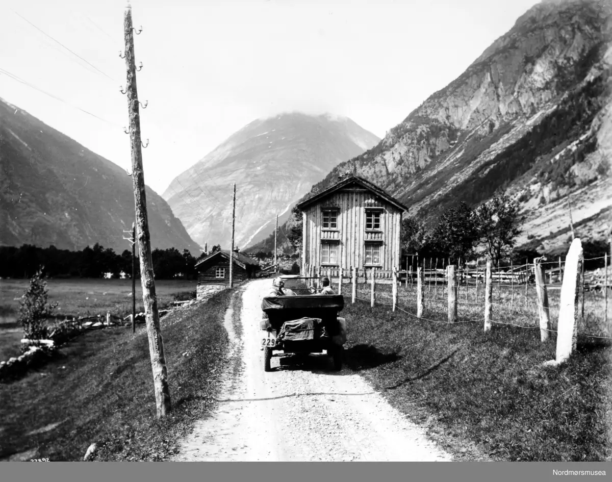 Foto nr. 22892 fra Anders Beer Wilses fotoarkiv, hvor vi ser fra en landevei ved Sunndalsøra. Bilen har registreringsskiltet "T-229". Fotoet er trolig fra perioden rundt 1910-1930. I følge Oddbjørn Skjørsæters notater (Romsdalsmseet) om rutebilene i Sunndalen ble T-229 brukt på en Chandler  7-seter 1919-modell. Bilen gikk i rutetrafikk for A/S Sundal-Opdal Auto, et selskap som skiftet navn til Kristiansund-Oppdal Auto A/S i 1932. Bilen gikk i rutetrafikk gjennom Sunndalen, mellom Sunndalsøra og Oppdal. (Info: Sveinung Berild - 2016). - Chandleren står i Bilboken for Norge 1922 (som 55/60 hk - kraftig for sin tid) og 1925, men nummeret er ikke i bruk i 1930-utgaven. (Info: Ivar Stav 2018 ).   Fra Nordmøre Museums fotosamlinger.