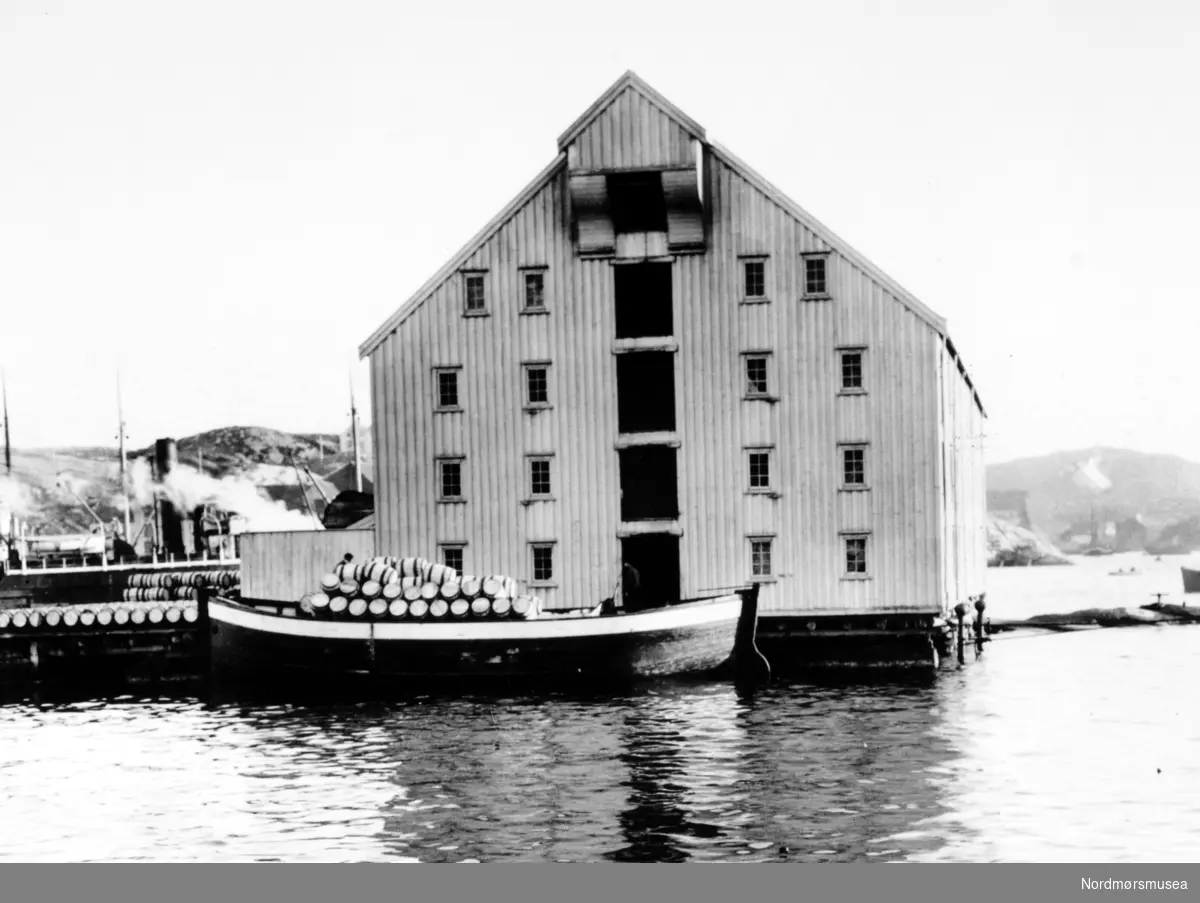 Foto nr. 25734 fra Anders Beer Wilses fotoarkiv, hvor vi ser fra ei brygge i Kristiansund kommune. Her er man igang med å laste en jekt(?) full av tønner,  trolig med saltet sild. Fotoet er trolig fra perioden rundt 1910-1930. Fra Nordmøre Museums fotosamlinger. 
