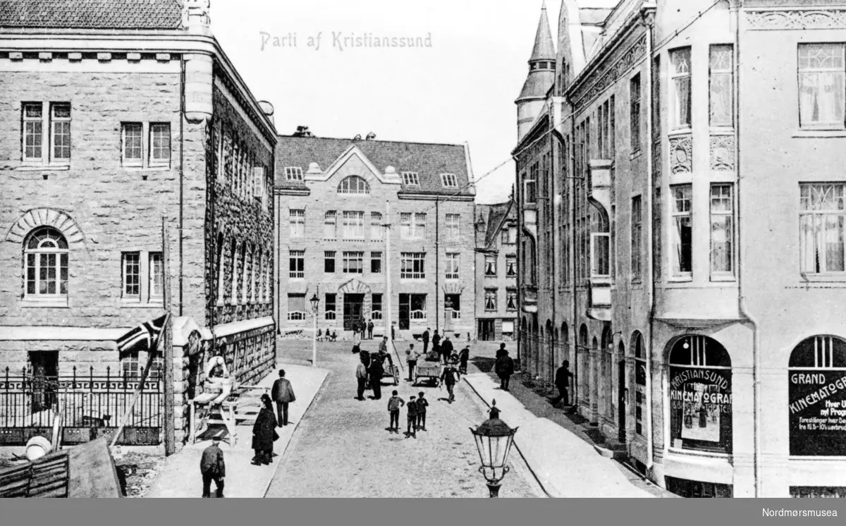 tønne, håndkjerre, Grand kino. Foto fotografert fra Øvre gade, med gaten Bernstorffstredet som går mellom Norges Banks bygning til venstre og Grand Hotell til høyre. Gaten leder inn til Torget, og vi ser i bakgrunnen kommunebygningen og noe av Gl&uuml;ckstadgården. Ellers så ser vi at Kristiansunds Kinematograf holder til i Grand hotell. Utgiver av prospektkortet er Georg R. Øyens papirforhandler. bank, kommunebygning, bygårder,

 Fra Nordmøre Museums fotosamlinger.
