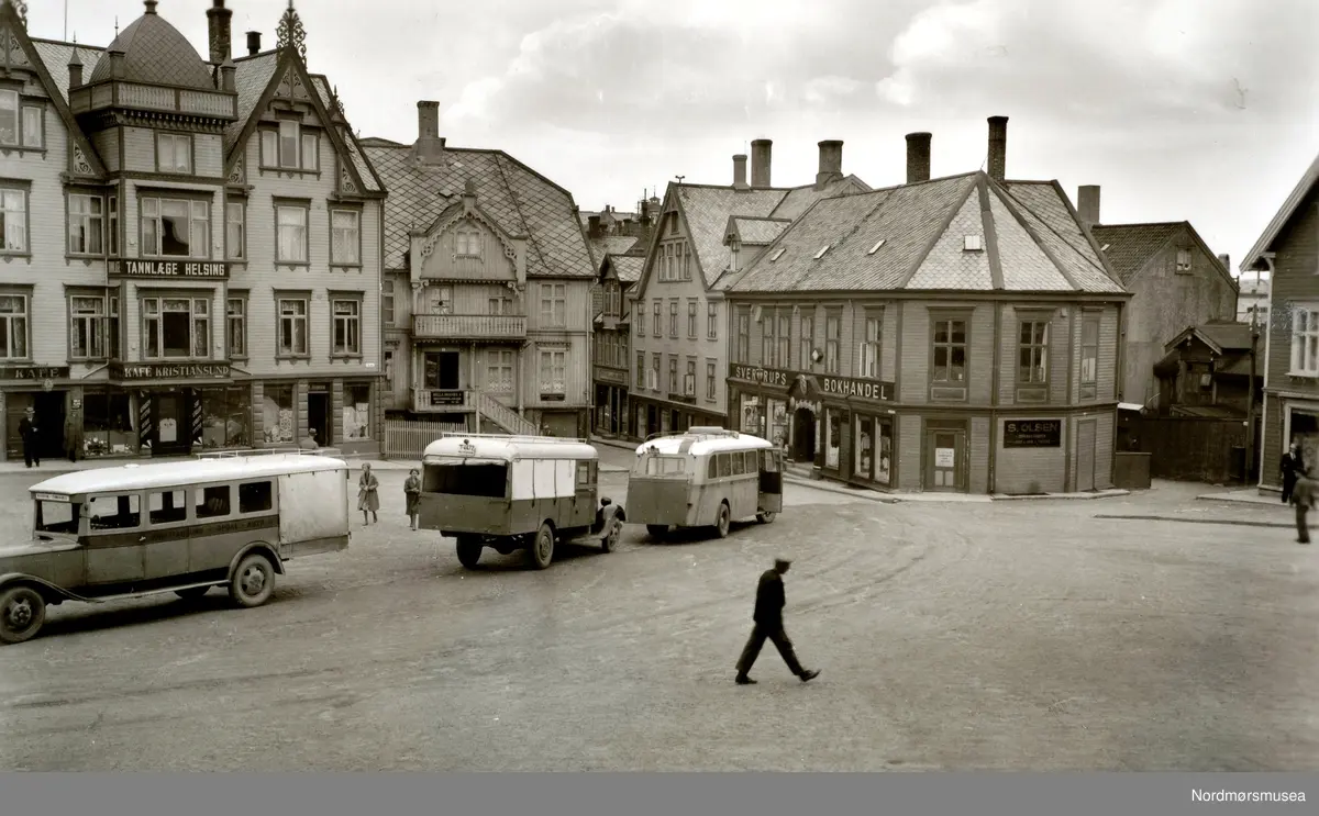 På bilen til venstre kan vi skimte Kristiansund-Oppdal Auto (KOA) på stripa under vinduene. Her er Oppdal stavet med P. Dette er nok av de tidlige rutebilene fra KOA. I 1932 åpnet ferjesambandet Kristiansund-Kvisvik. KOA og Surnadal Billag sine rutebiler kunne dermed følge ferja til Kristiansund sentrum. Det var først i 1938 at rutebilene fra Molde og Gjemnes kunne følge ferja til Kristiansund. Det var dette året ferjesambandet til Gjemnes og Torvikbukt åpnet. Bildet er derfor tidligst fra 1932. (Info: Sveinung Berild - 11.11.2016). - Postkort med motiv fra Torvet på Kirkelandet i Kristiansund. Her står busser klar for transport. Til høyre ser vi Sverdrups Bokhandel som også er utgiver at dette kortet i 1936. bil, transport, butikk, bokhandel, personer

 Fra Nordmøre Museums fotosamlinger.