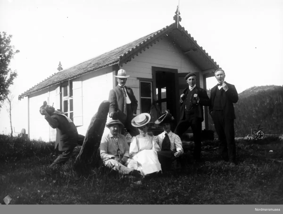 Foto av en gruppe menn og kvinner, trolig på landtur, med en hytte like bak. Det er ukjent hvem vi ser på bildet, samt hvor og når bildet er tatt. Fra Nordmøre museums fotosamlinger.
