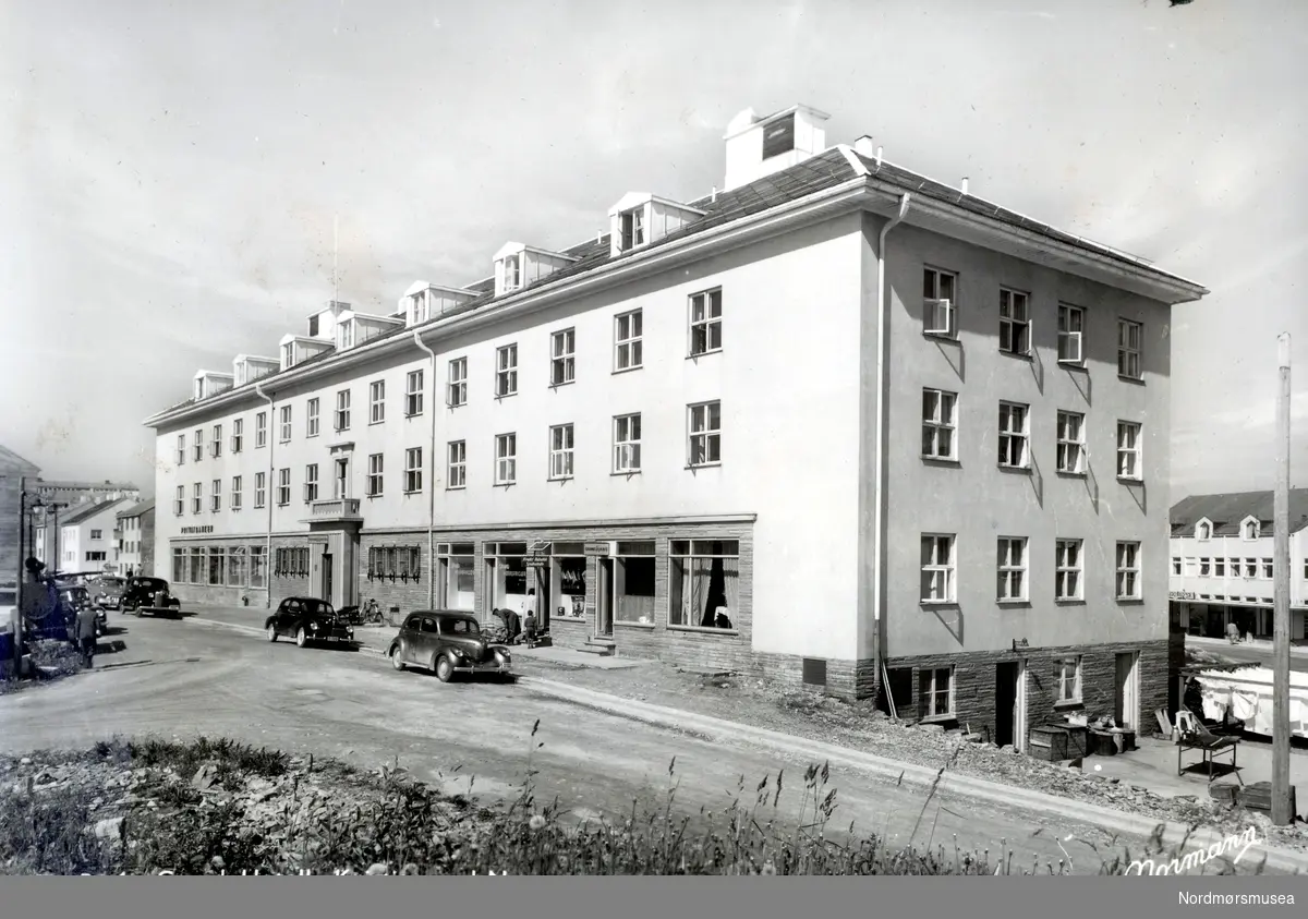 Jan Messel Williamsen, f. 1949, skriver: Grand Hotell ca 1955. Utsnitt fra et postkort fra Normann. Fotografen stod trolig oppå bunkersen som senere ble revet til fordel for Storgata 18. Dette er fra reviret der jeg vokste opp. Forretninger i Bernstorfftstredet: Fra høyre ser vi Grand Cafe. Her var det ølservering. Mørelinjen og Oppdal Auto hadde stopp her. Av og til ble det et par øl for mye på dem som ventet på bussen. Neste dør der en mor og en gutt er på vei inn var Harald Bergsli Frukt og Tobakk. Der fikk vi småguttene kjøpe drops for 2 øre stykket. Alle guttene i gata kjente Harald. Han solgte også ukeblader og den smale utgaven av Wild West med kaptein Miki, Windy og Salasso. Harald var tidligere sjømann og bokser. En gang spilte han bokser i en film som gikk på kino. Alle guttene fra Storgata-Knudtzonsgata og bortover til Langveien og Ramsaygata var på kino og så filmen. Senere drev Bergsli kiosken på Piren. Mannen med lagerfrakk og sykkelvogn var trolig vaktmester på Grand. Butikken bortenfor ham var Grand Delikatesse. Der kunne man kjøpe varm middag og ta med hjem. Lengre bort er hovedinngangen til Grand, Resepsjon står det på skiltet. Det gikk an å kjøpe frimerke i resepsjonen. Og ville man ha rask postgang eksempelvis til Bergen, så gikk man ned på kaia og postet på Hurtigruta. Den la til utenfor Nordmøre Meieri og Brødrene Dall. Man måtte se om det var sørgående, ellers ble det sen postgang om Kirkenes. Utenfor står også en gammel dragkjerre av tre med jernringer på hjulene. En sykkel står ved siden av. Uti gata står bakerst en Peugeot 203, den ble produsert fra 1948 til 1960. Foran står en Willys, sannsynlig en 1939 modell. 

Postkort med tekst:  ";Grand Hotell,  Kristiansund N.";  (Fra Nordmøre Museums fotosamlinger. Dublett Kmb-1987-005.0169, fra nyere: KMb-2016-006.0008