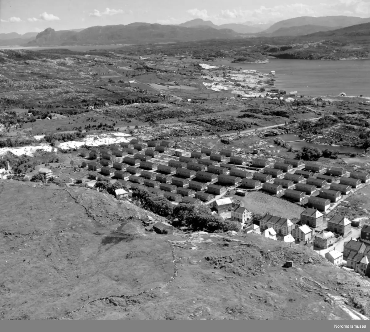 Brakkebyen på Nordlandet fotografert i fra Bjønnahaugen. En del av barakkene som ble oppsatt for erstatning for de bombede og nedbrente boligene under bombingen av byen i april 1940. Nedenfor haugen ser en bebyggelsen fra 1915-20 årene og gårdsbruket til Boksaspen. Til høyre ser en bebyggelsen i Vikan og Bolgsvaet. Lengst til høyre ligger Rensvik på Frei.
I bakgrunnen
ser vi
fjellene på Straumsnes med Valfjellet, Årsundfjellet, Strandafjellet, Liafjellet og det karakteristiske Magnhildberget. (Tilleggsinfo: Ronny Bolgen.)



