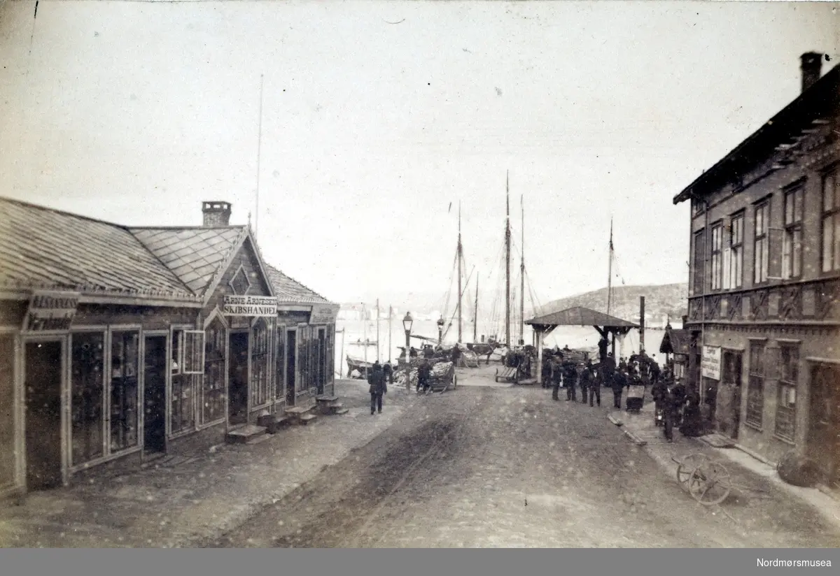 "Kirkealmenningen". Foto fra deler av bebyggelsen i Kirkeallmenningen på Kirkelandet i Kristiansund. Her ser vi blant annet Arne Arnesens Skipshandel til venstre på bildet, men Allmenningen i enden av gaten. Bildet er fra fotoalbumet "Christiansund i Fotografeir". Bildet er trolig fra 1883 etter informasjon fra Willhelm Lund samlingen. Fra Nordmøre Museums fotosamlinger. dublett, ett av mange