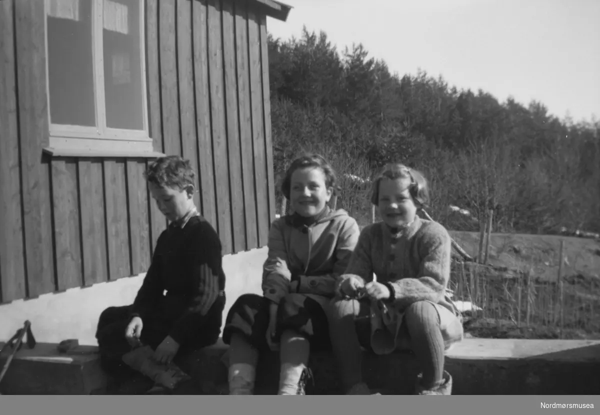 Foto av tre barn - en gutt og to piker - sittende utenfor et bolighus. Trolig enten familie, slekt eller bekjente av fotografen, som sannsynligvis er John Myren. Datering er ukjent, men trolig mellom 1930 til 1960. Stedet er også ukjent, men kan være fra Sunndal kommune. Fra Nordmøre museums fotosamlinger.

