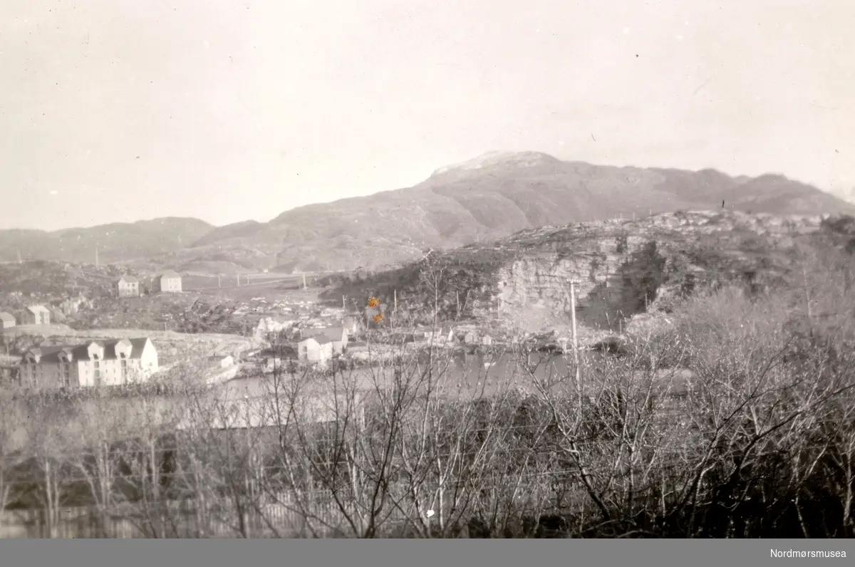 Fotografen har stått i sykehusets hage og fotografert mot Innlandet hvor vi ser steinbrudet der de tok stein i 1920-30 tallet (nødsarbeid) for bygging av blant annet moloen i Sørsundet. Det hadde lenge vært gjort krav om at havneforholdene måtte forbedres, spesielt etter at dampskipet ";Norden"; kom i havsnød, sleit fortøyningene og havnet opp i fjæra her i Lervika den12.02.1903. Se Kmb-2008-026.0018 og Kmb-2008-026.0043. Til venstre for steinbruddet kommer i dag, 2009, Sørsundbrua fra Kirkelandet til Innlandet, som i 1963 fikk fast forbindelse med med resten av Kristiansund. Den lille husklyngen ligger i dag under brua. Til venstre ligger ei brygge i Lervika og noen av husene i Kaptein Bothners gate. Videre til venstre ser vi de to store bryggene til Backer og Mikkelsen. I høyden bak den kvite Backerbrygga ble to hus oppført før 1. verdenskrig og her ble informanten Leif Rødsand født i huset til høyre. Det var fire leiligheter i hvert av husene. Foruten familien Rødsand bodde også familiene Wåge, Olsen og ? der. I det andre huset bodde Bævre som var eier og Sørlie, han som mistet handa, og ? ?. Månedsleien var kr. 30. Da det ble ledig leilighet i Heinsagata til kr. 25, flyttet familien Rødsand dit. 5 kroner var mange penger i den tiden. Nedenfor mot venstre ser vi litt av husene i Reknergata. Den nye innfartsveien fra brua passerer Reknergata ned mot Heinsagata, Garverigata og videre mot Fiskergata, Beatebakken, Skippergata og Skjærvaveien. Innlandet ble lite ødelagt i forbindelse bombingen av Kristiansund i aprildagene 1940 i forhold til de andre ";landene";, Kirkelandet, Gomalandet og Nordlandet. Det var bebyggelsen langs Markussundet som ble bombet på Innlandet. I bakgrunnen ser vi Freikollen som tilhørte nabokommunen Frei, men som fra 1. januar 2008 ble innlemmet i Kristiansund. Fra Ida M. Knudtzons fotosamlinger, nå en del av Nordmøre Museums fotosamlinger. serie
