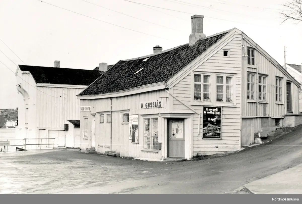 Foto fra landhandelen til Magnar Gussiås fra Dødeladen på Innlandet i Kristiansund. Dødeladen er opprinnelig naustet til Tollboden, den store staselige brygga som ligger vegg i vegg. Tollboden er fra ca 1660, og var det økonomiske senteret i kjøpstaden. Her ble alle varer ble registrert og fortollet. Tollbod-funksjonen lå her fram til 1742, da den ble flyttet over til Kirkelandet. Brygga er nå under restaurering.

Dødeladen ble bygd som naust ca 1700, og er seiner ombygd og utbygd i alle retninger. Har var kanskje byens første rådhus, og bygningen har huset alt fra sprit og vin utsalg, bakeri, kolonialbutikk til søndagsskole! Her har det vært boksetreningslokale (for den svært lokale klubben ";Treff"; , skomaker og bolig blant mye annet. Navnet Dødeladen kommer av en forsikrings-ordning som hadde kontor i 2 etg. hvor spesielt fiskere og sjøfolk, -eller andre, betalte noen øre hver måned til sin egen begravelse!

Drengstua som hørte til eiendommen ligger her enda, og er nå restaurert og brukes til møtelokale. I tillegg har Tahitibrygga kommet til konseptet. Dette var ei salt-brygge, bygd ca 1900. Her er det nå selskapslokaler/kulturlokaler som drives av Dødeladen/Vikaneset.

Merk at reklameplakaten ikke er den samme som på KMb-2008-052.0032.

Før Dødeladen restaurant drev ei dame kafé;en Plutos Verdenshjørne her et par år. Frosne boller i mikroovn.


Fra Nordmøre museums fotosamlinger.



