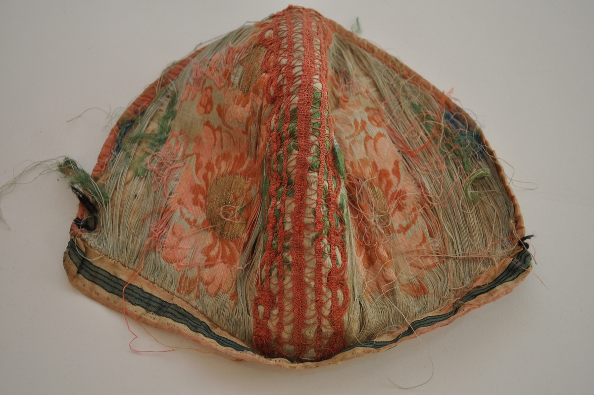 Tobladlue i lys, blågrønn silkesateng med innvevde, brosjerte blomstermotiver i lyserødt, rødbrunt, grønt og blått samt gulltråd. Stoffet kan dermed karakteriseres som brokade. Over midtsømmen løper en lakserød blonde og langs nederkanten er et stripet silkeband i lyserødt, hvitt, grønt og svart. Framkanten er kantet med et lyserødt silkeband. Fôret er av ubleiket linlerret. Lua er sydd sammen med silketråd i hvitt, blått og lyserødt. Silkestoffet er brustent, så lua framstår som den er har dårlig tilstand.