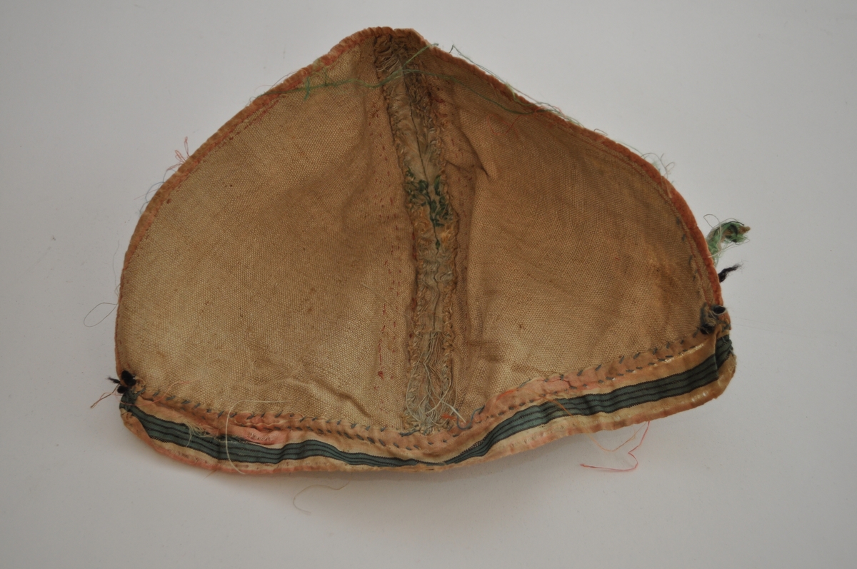 Tobladlue i lys, blågrønn silkesateng med innvevde, brosjerte blomstermotiver i lyserødt, rødbrunt, grønt og blått samt gulltråd. Stoffet kan dermed karakteriseres som brokade. Over midtsømmen løper en lakserød blonde og langs nederkanten er et stripet silkeband i lyserødt, hvitt, grønt og svart. Framkanten er kantet med et lyserødt silkeband. Fôret er av ubleiket linlerret. Lua er sydd sammen med silketråd i hvitt, blått og lyserødt. Silkestoffet er brustent, så lua framstår som den er har dårlig tilstand.