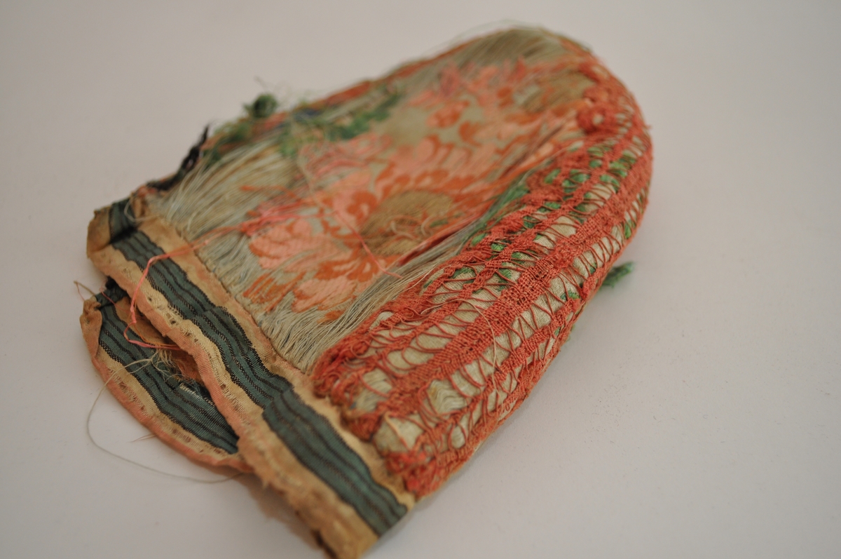 Tobladlue i lys, blågrønn silkesateng med innvevde, brosjerte blomstermotiver i lyserødt, rødbrunt, grønt og blått samt gulltråd. Stoffet kan dermed karakteriseres som brokade. Over midtsømmen løper en lakserød blonde og langs nederkanten er et stripet silkeband i lyserødt, hvitt, grønt og svart. Framkanten er kantet med et lyserødt silkeband. Fôret er av ubleiket linlerret. Lua er sydd sammen med silketråd i hvitt, blått og lyserødt. Silkestoffet er brustent, så lua framstår som den er har dårlig tilstand.