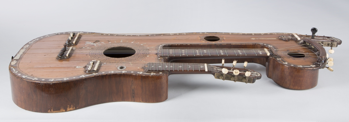 Trippelgitar, 3 halser. Lokk av gran, består av 3 deler, bunn og sarg muligens en sort mahogny. Den lengste halsen fortsetter øverst ut i en ny resonanskasse, hvis overside tjener som skruekasse for den mellomste halsen. Her er også et lydhull. En ekstra resonanseffekt oppstår ved at strengene som løper over denne resonanskassedelen under spillet svinger med. Instrumentet er utsmykket med rike perlemosrsforsiringer, delvis stekt fargede. Ornamentene er dels svært enkle og dels meget delikate blomstermønstre. Hovedlydhullet er omgitt av en 4 cm. bred krans av geometrisk intarsia, som kan minne om maurisk stil. Instrumentet har i alt 4 lydhull og 3 adskilte strengestoler. Den lengste halsen har 7 frittsvingende bass-strenger de to andre hver 6 strenger og 13 tverrbånd av metall. Runde skruehoder av elfenben. Likeså oversadler og strengestoler av elfenben.
Usignert.