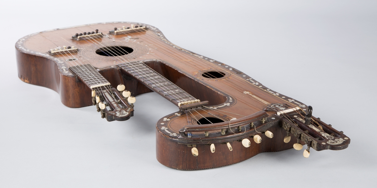 Trippelgitar, 3 halser. Lokk av gran, består av 3 deler, bunn og sarg muligens en sort mahogny. Den lengste halsen fortsetter øverst ut i en ny resonanskasse, hvis overside tjener som skruekasse for den mellomste halsen. Her er også et lydhull. En ekstra resonanseffekt oppstår ved at strengene som løper over denne resonanskassedelen under spillet svinger med. Instrumentet er utsmykket med rike perlemosrsforsiringer, delvis stekt fargede. Ornamentene er dels svært enkle og dels meget delikate blomstermønstre. Hovedlydhullet er omgitt av en 4 cm. bred krans av geometrisk intarsia, som kan minne om maurisk stil. Instrumentet har i alt 4 lydhull og 3 adskilte strengestoler. Den lengste halsen har 7 frittsvingende bass-strenger de to andre hver 6 strenger og 13 tverrbånd av metall. Runde skruehoder av elfenben. Likeså oversadler og strengestoler av elfenben.
Usignert.