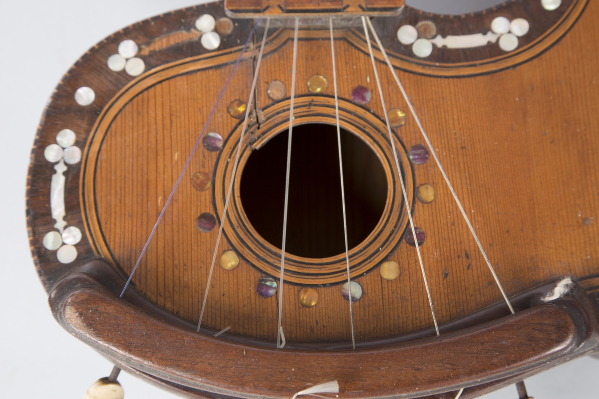 Trippelgitar, 3 halser. Lokk av gran, består av 3 deler, bunn og sarg muligens en sort mahogny. Den lengste halsen fortsetter øverst ut i en ny resonanskasse, hvis overside tjener som skruekasse for den mellomste halsen. Her er også et lydhull. En ekstra resonanseffekt oppstår ved at strengene som løper over denne resonanskassedelen under spillet svinger med. Instrumentet er utsmykket med rike perlemosrsforsiringer, delvis stekt fargede. Ornamentene er dels svært enkle og dels meget delikate blomstermønstre. Hovedlydhullet er omgitt av en 4 cm. bred krans av geometrisk intarsia, som kan minne om maurisk stil. Instrumentet har i alt 4 lydhull og 3 adskilte strengestoler. Den lengste halsen har 7 frittsvingende bass-strenger de to andre hver 6 strenger og 13 tverrbånd av metall. Runde skruehoder av elfenben. Likeså oversadler og strengestoler av elfenben.
Usignert.