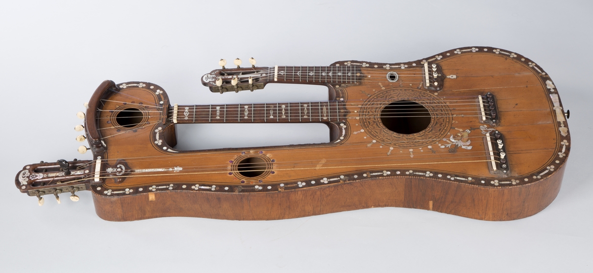 Trippelgitar, 3 halser. Lokk av gran, består av 3 deler, bunn og sarg muligens en sort mahogny. Den lengste halsen fortsetter øverst ut i en ny resonanskasse, hvis overside tjener som skruekasse for den mellomste halsen. Her er også et lydhull. En ekstra resonanseffekt oppstår ved at strengene som løper over denne resonanskassedelen under spillet svinger med. Instrumentet er utsmykket med rike perlemosrsforsiringer, delvis stekt fargede. Ornamentene er dels svært enkle og dels meget delikate blomstermønstre. Hovedlydhullet er omgitt av en 4 cm. bred krans av geometrisk intarsia, som kan minne om maurisk stil. Instrumentet har i alt 4 lydhull og 3 adskilte strengestoler. Den lengste halsen har 7 frittsvingende bass-strenger de to andre hver 6 strenger og 13 tverrbånd av metall. Runde skruehoder av elfenben. Likeså oversadler og strengestoler av elfenben.
Usignert.