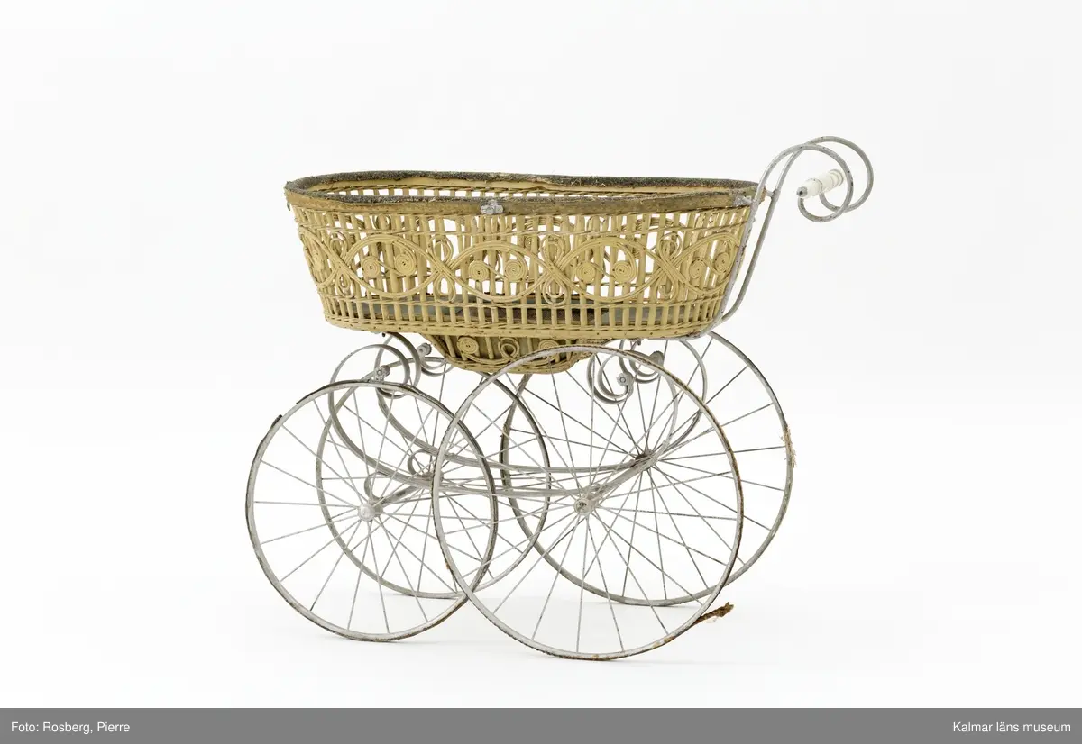 KLM 28338:1. Barnvagn, liggvagn. Hjul och underrede av metall målade i silverfärg, troligen ej ursprunglig. Två hjul är gummiklädda, ett saknar detta och det fjärde är försett med flätat naturmaterial. Handtag av vitt porslin. Vagnskorg med trästomme bestående av flätade stående och liggande spjälor målade i ljusgul nyans. Övre kanten av korgen är klädd med grovt ylletyg och på utsidan ett vävt tygband i gul ylle. Korgens botten är klädd med grönt konstläder och säckväv målat i rutmönster. I mitten av korgen en försänkning. På korgens sidor fästen för sufflett. På hjulen instansat: BRENNABOR D.R.G.M.