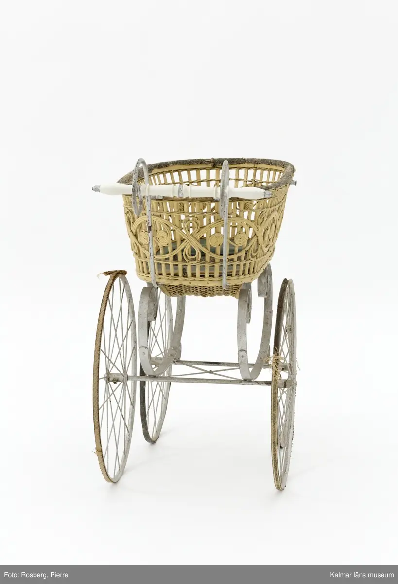 KLM 28338:1. Barnvagn, liggvagn. Hjul och underrede av metall målade i silverfärg, troligen ej ursprunglig. Två hjul är gummiklädda, ett saknar detta och det fjärde är försett med flätat naturmaterial. Handtag av vitt porslin. Vagnskorg med trästomme bestående av flätade stående och liggande spjälor målade i ljusgul nyans. Övre kanten av korgen är klädd med grovt ylletyg och på utsidan ett vävt tygband i gul ylle. Korgens botten är klädd med grönt konstläder och säckväv målat i rutmönster. I mitten av korgen en försänkning. På korgens sidor fästen för sufflett. På hjulen instansat: BRENNABOR D.R.G.M.