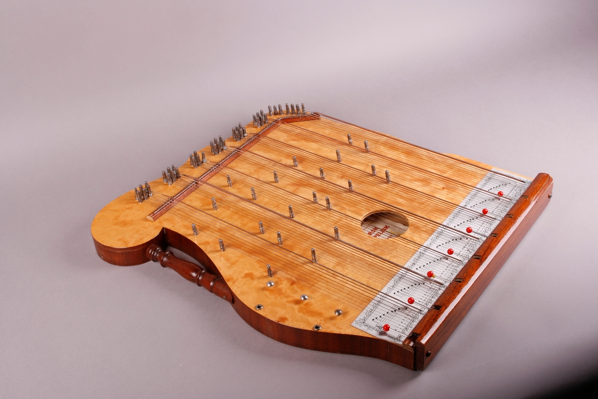 Rektangulær korpus med lokk og bunn i lysebrunt tre. Sargen er mørkt tre. Bunnen er flat. Nedre sarg er flat, og de andre tre sidene er buet. Instrmentet har en "arm" på venstre side. Fra denne armen går et håndtak i form av en søyle. Tre metallknapper på venstre side nedenfor håndtaket. Ved disse er det to metallskruer/ringer her som lager hull inn i resonanskassen. 

64 strenger som er gruppert i sju akkorder. Fra venstre: F-dur, C-dur, G-dur, D-dur, A-dur, E-dur, H-dur. Nederst på instrumentet er det påmontert et brett som beskytter strengefestet. Under strengene går det for hver akkord en stang som kan bøyes oppover. Med disse kan man forandre tersen i hver akkord, og dermed spille i moll. 

Nederst, før strengefestet og brettet er det skrudd på en metallplate som beskriver tonehøyder via notelinjer, samt notenavn. Etikett på kremhvitt (falmet?) papir er pålimt inne i kassen.