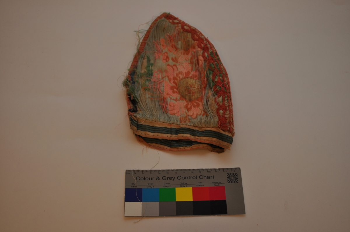 Tobladlue i lys, blågrønn silkesateng med innvevde, brosjerte blomstermotiver i lyserødt, rødbrunt, grønt og blått samt gulltråd. Stoffet kan dermed karakteriseres som brokade. Over midtsømmen løper en lakserød blonde og langs nederkanten er et stripet silkeband i lyserødt, hvitt, grønt og svart. Framkanten er kantet med et lyserødt silkeband. Fôret er av ubleiket linlerret. Lua er sydd sammen med silketråd i hvitt, blått og lyserødt. Silkestoffet er brustent, så lua framstår som den er har dårlig tilstand.