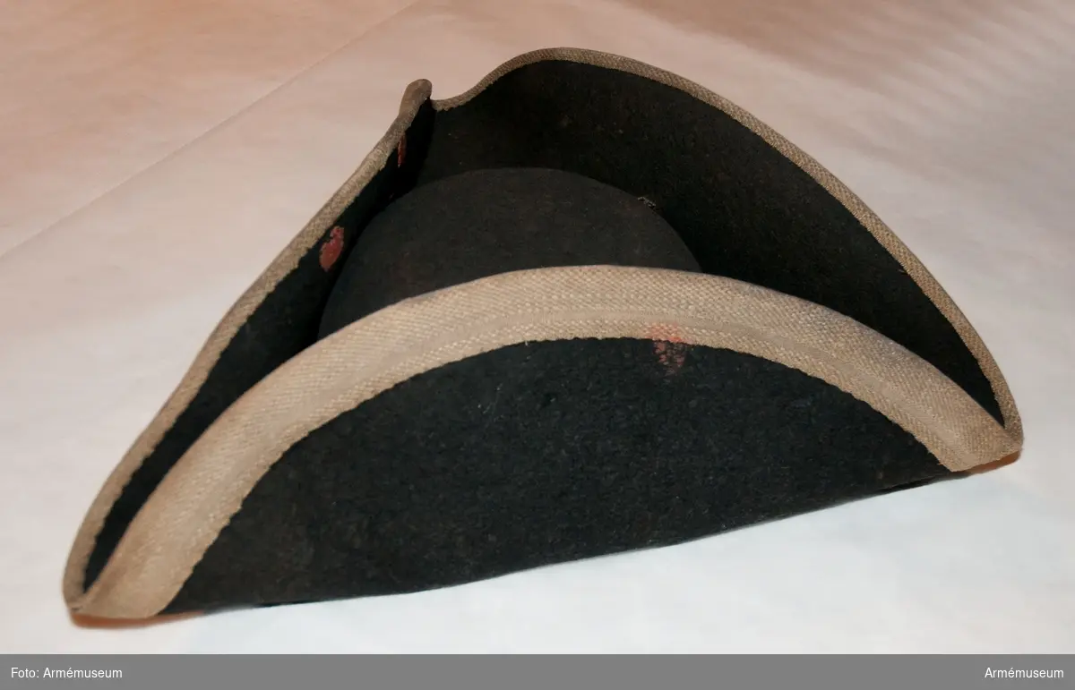 Trekantig hatt av svart filt med vitt band utmed brättet.
