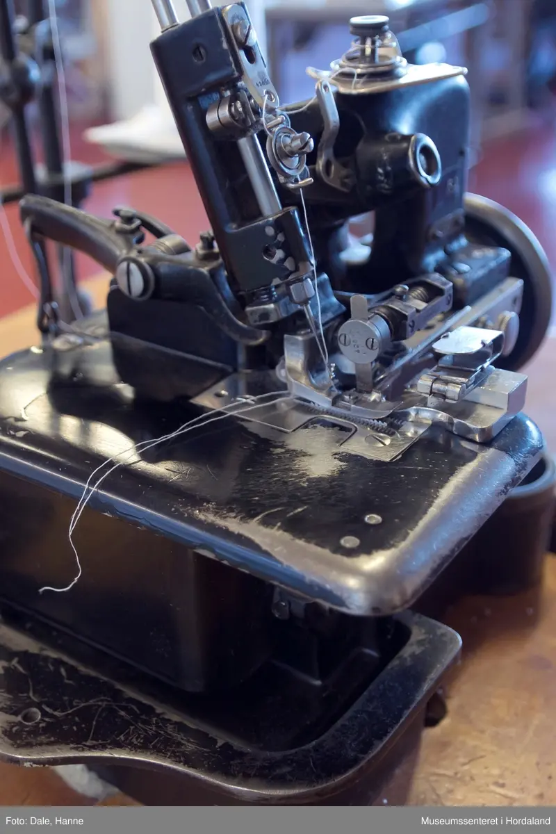 Ei to-tråds Singer symaskin til falding/kantar på strikka stoff, med original motor, lampe, montert på bord. 

Modell 81-6, merka med nummer 54. Maskinnummer AB503010. Overlockmaskin med faldeapparat, til kanting av stoff.