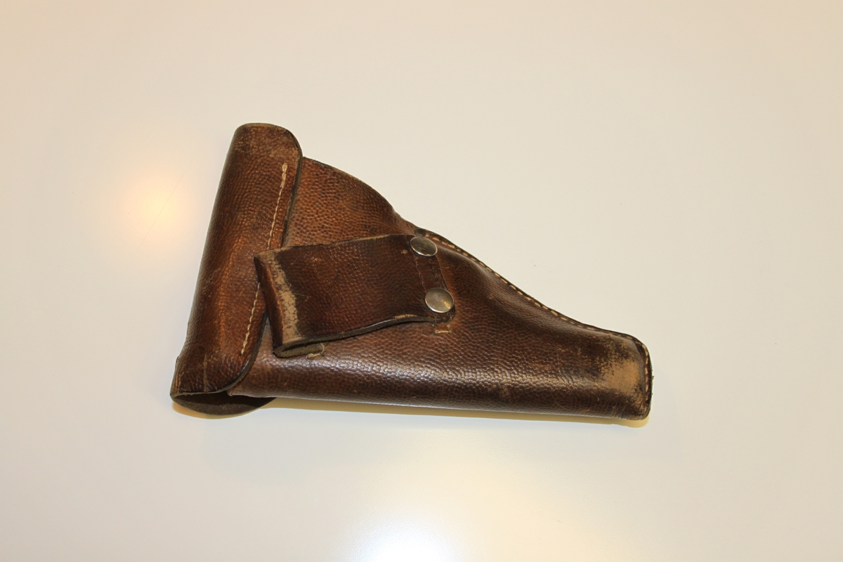Hylse i brunt lær med lukning høyre side. Lukker ved hjelp av rund knapp i spor. Sydd.

Egen lomme høyre side til ekstra magasin. Ekstra magasin ligger i. 

Hylse har stempel på innsiden av lokk: 
Vanskelig å lese men er bokstaver og tall. Ovalt stempel.

Venstre side har feste til belte. Beltet tres da gjennom feste. Sydd på i samme lær som resten.
